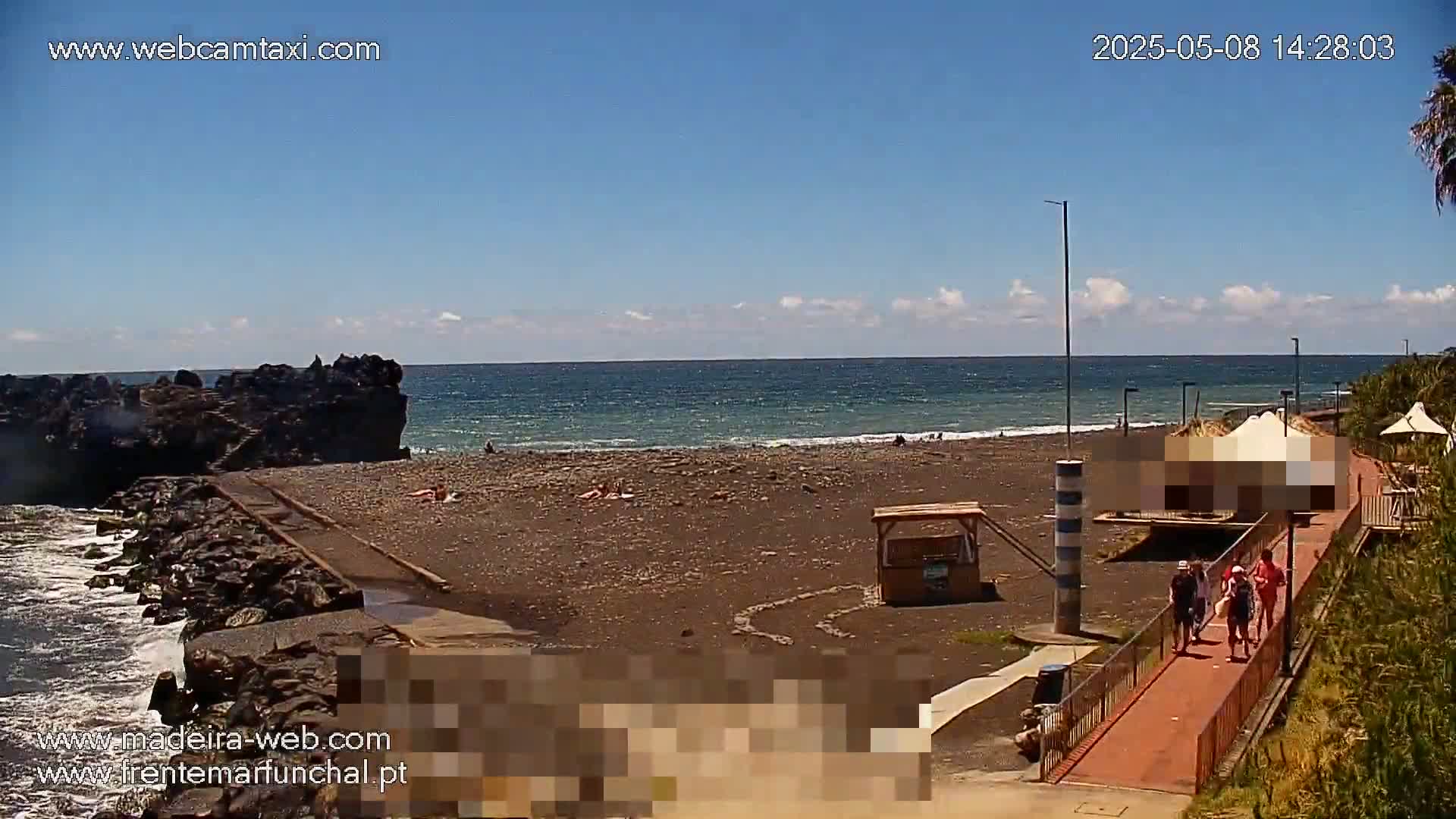 A dark-sand beach with a few people on it, near a rocky outcrop, stretches along a calm ocean under a mostly clear blue sky.