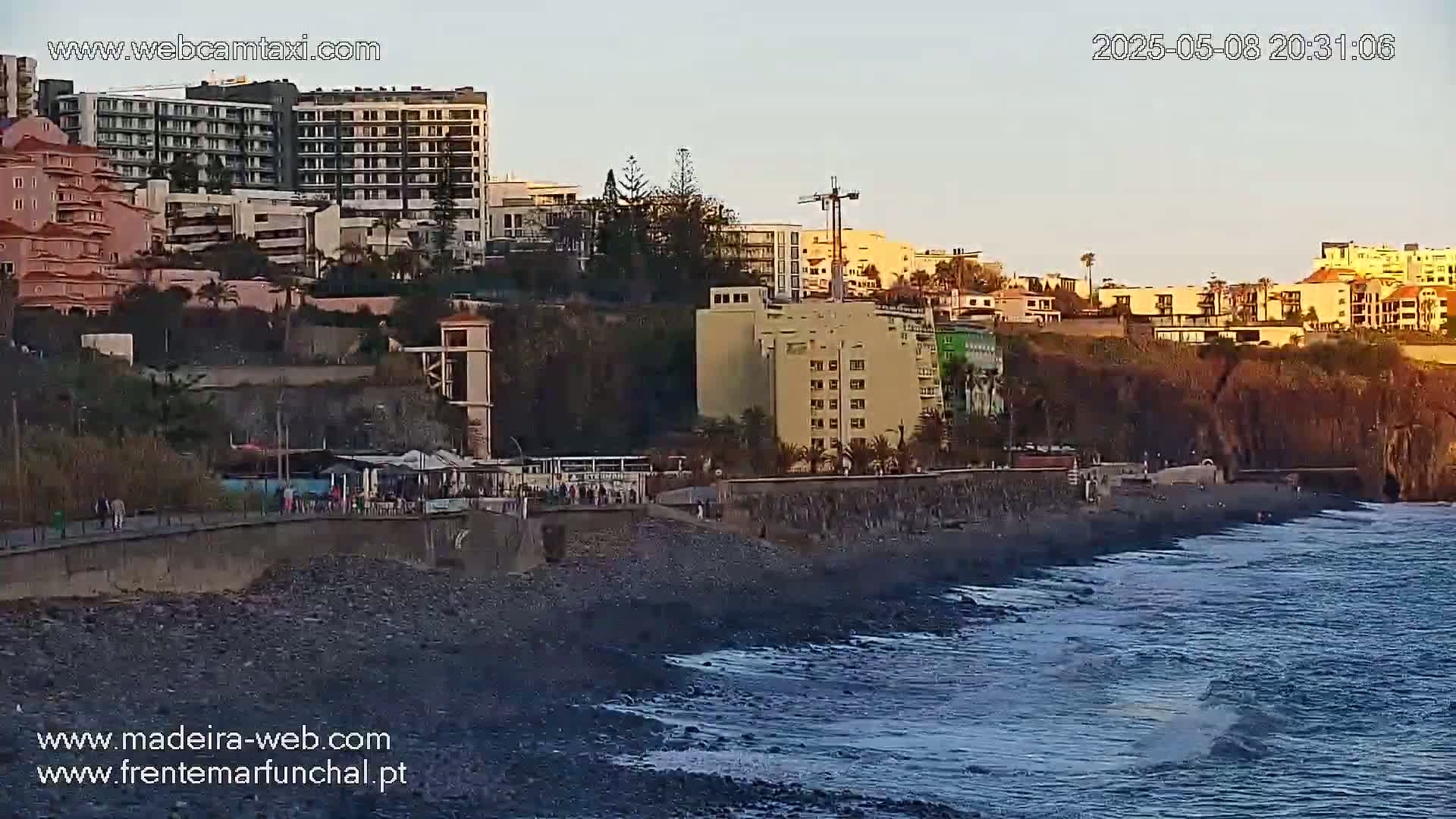 A rocky beach with gentle waves laps at the shore, under a twilight sky, near a collection of buildings and hotels on a cliffside.