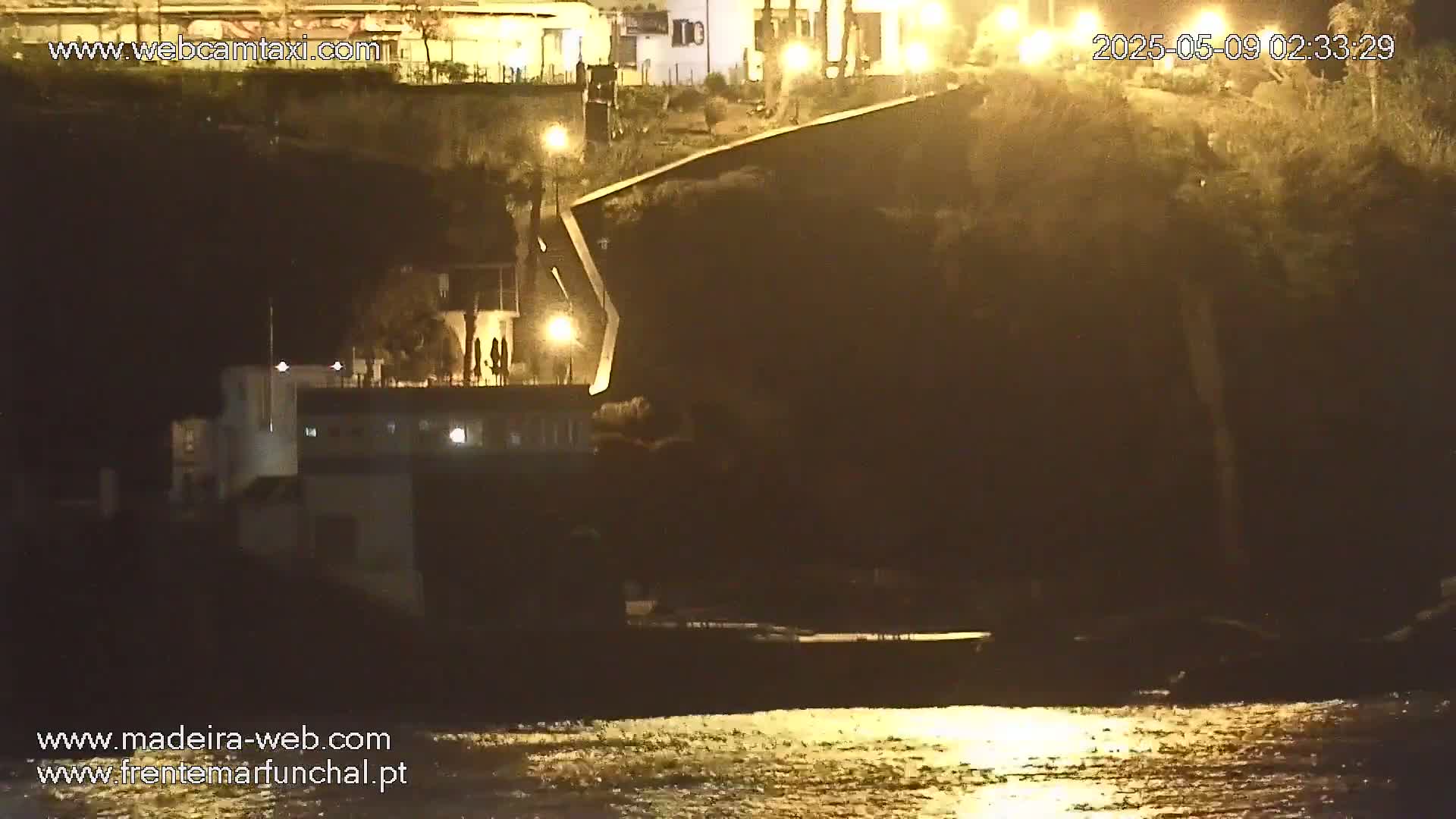 A nighttime waterfront scene shows a dark cliffside with buildings and lights, reflecting onto calm water.
