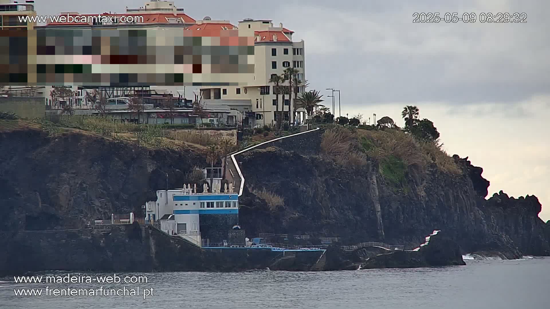 A multi-story building sits atop a dark cliff overlooking a body of water under an overcast sky.