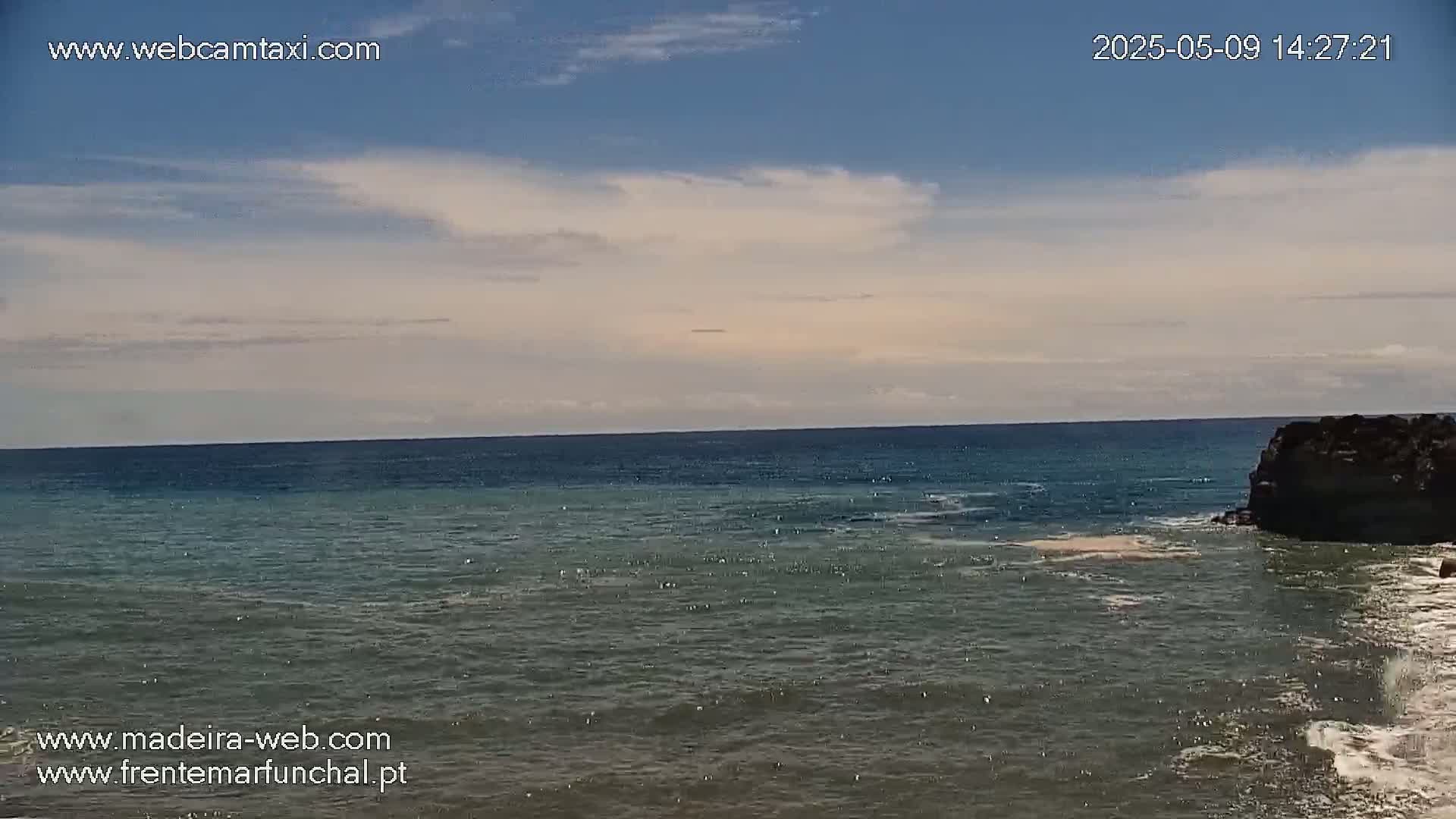 A mostly sunny day shows a calm ocean meeting a rocky shoreline under a partly cloudy sky.