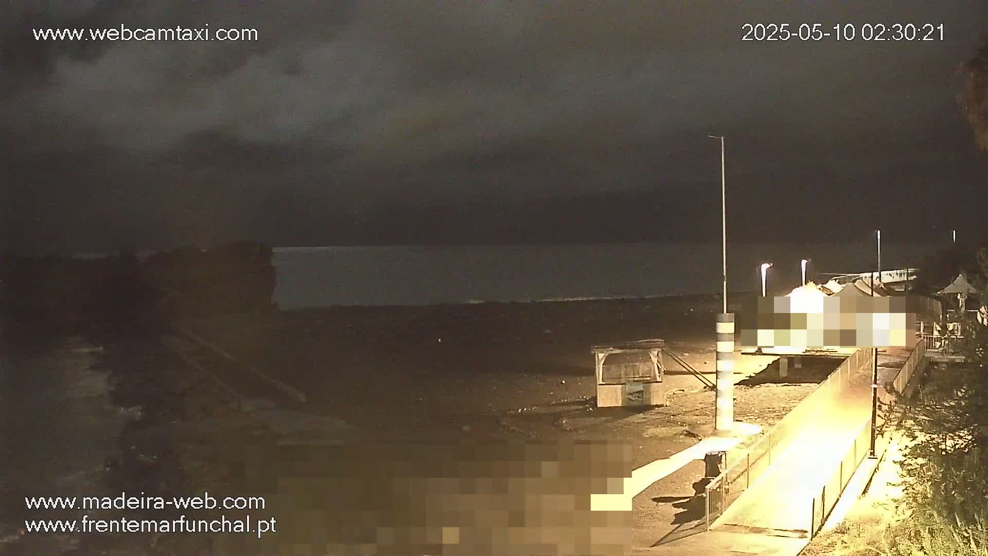 A nighttime view of a dark beach with a calm sea under an overcast sky shows a walkway leading to a structure near the water's edge.