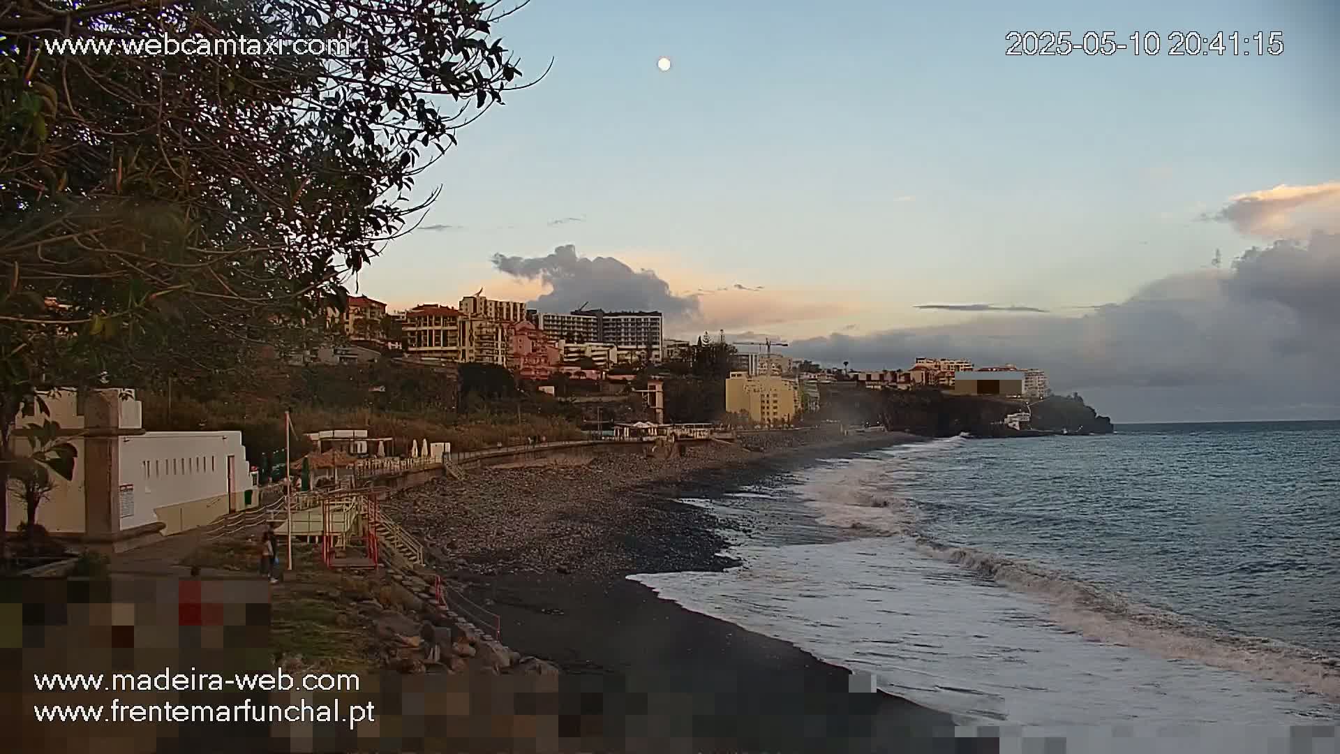 A dark-sand beach with gentle waves laps the shore under a partly cloudy twilight sky, with buildings and a moon visible in the distance.