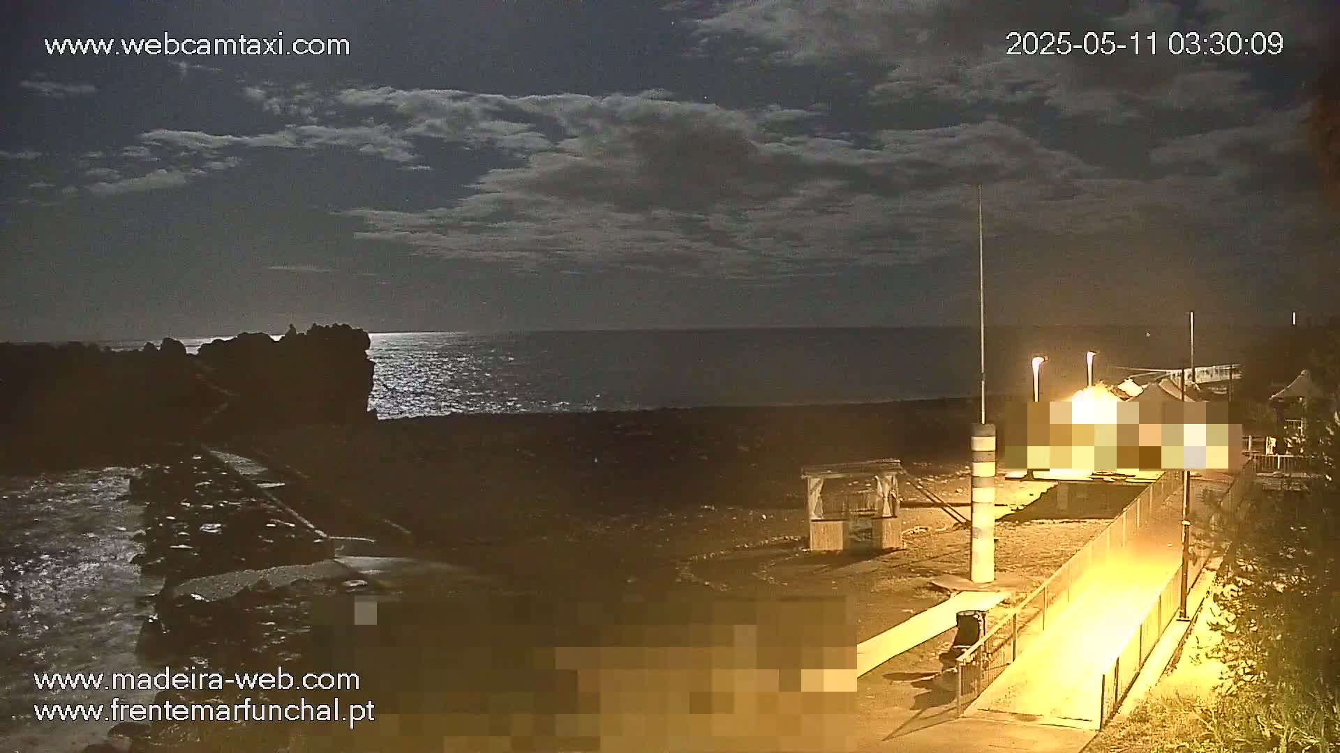 A nighttime beach scene under a partly cloudy sky shows a dark ocean, rocky shoreline, and illuminated buildings near a walkway.