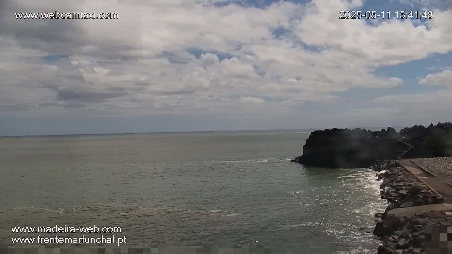 A partly cloudy sky overlooks a calm ocean meeting dark rocky shores with some small waves.
