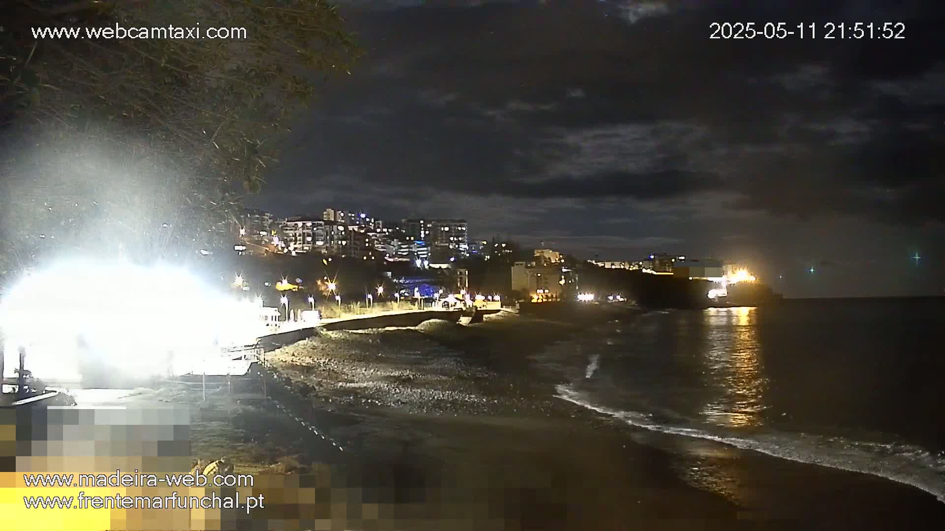 A nighttime coastal scene shows a dark beach with gentle waves lapping the shore under a cloudy sky, with illuminated buildings visible in the distance.