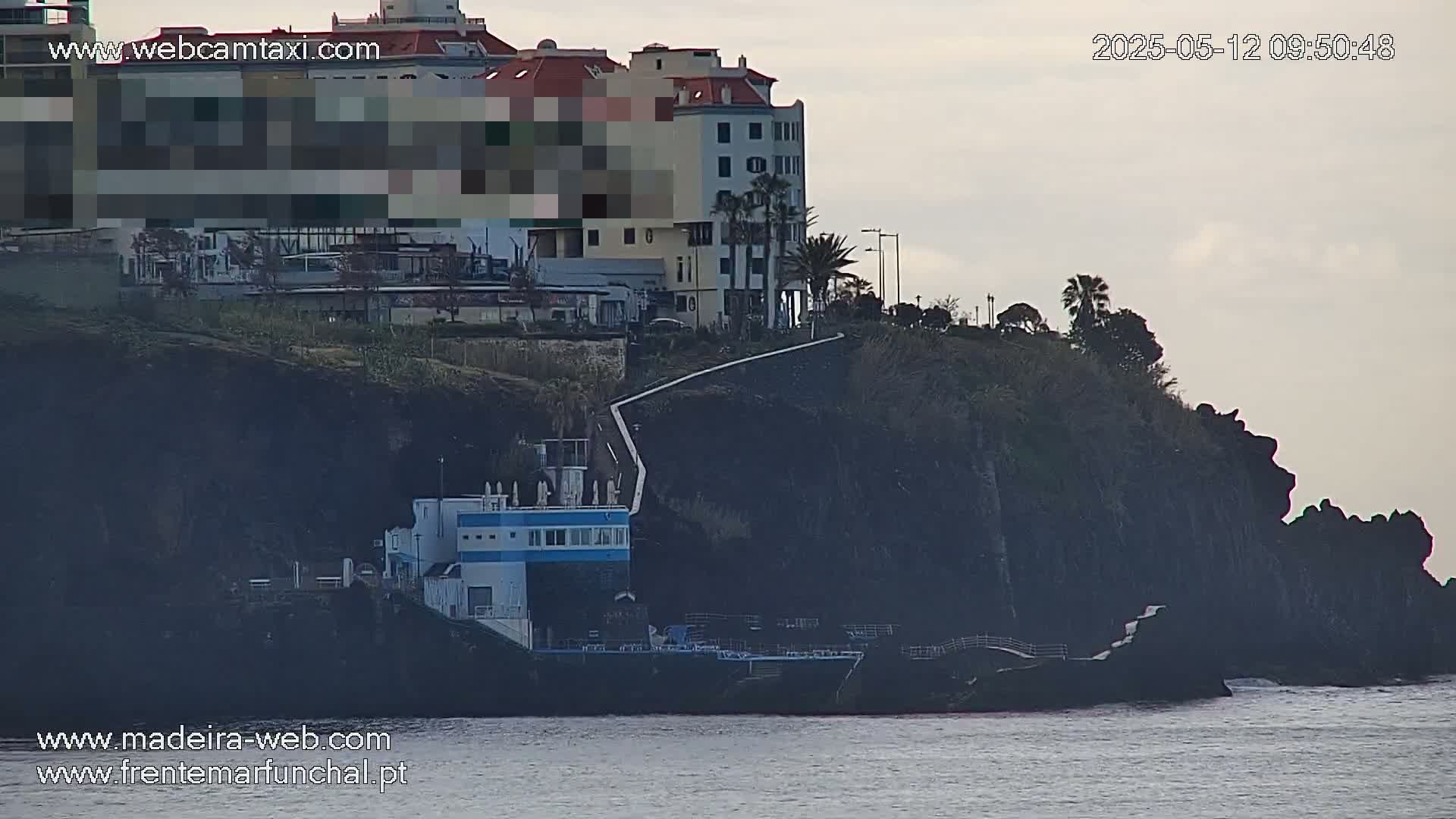 A coastal scene under an overcast sky shows a building perched atop dark cliffs overlooking a calm body of water.