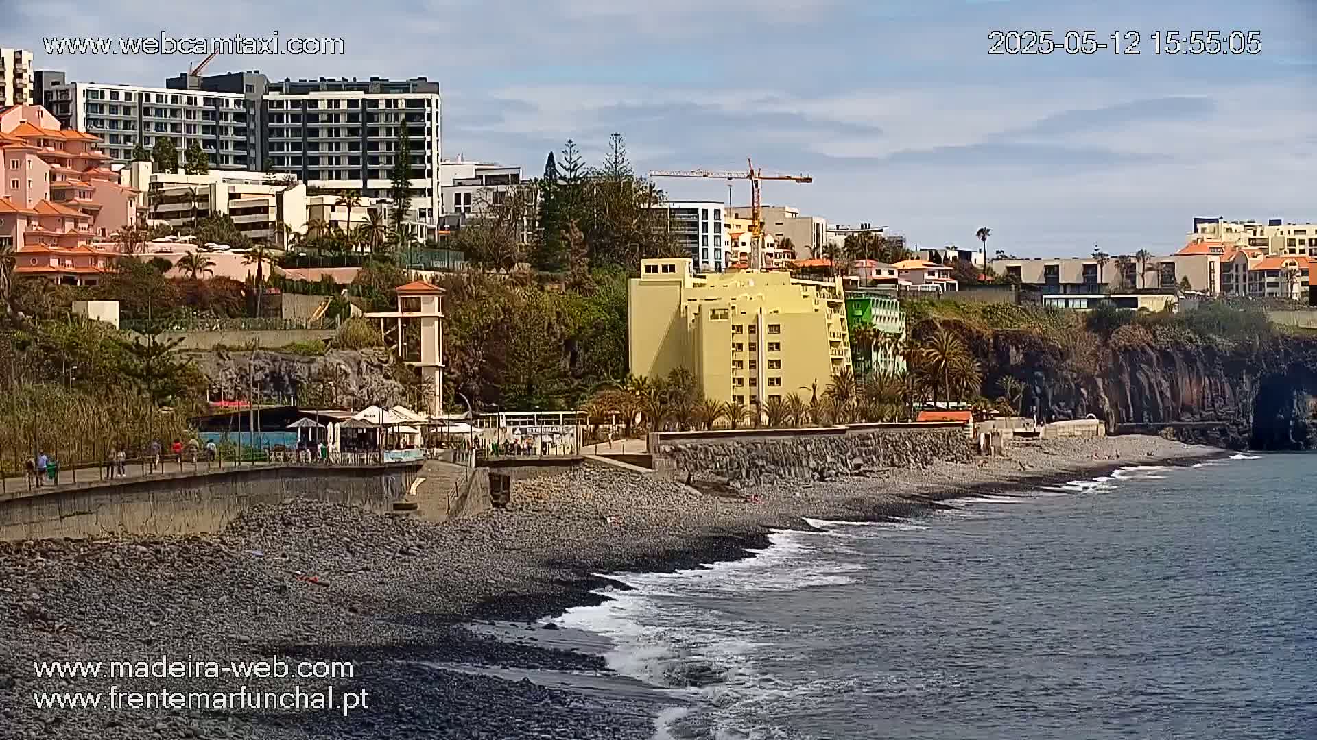 A rocky beach with a few people and buildings on a hillside overlooking calm ocean water under a partly cloudy sky.