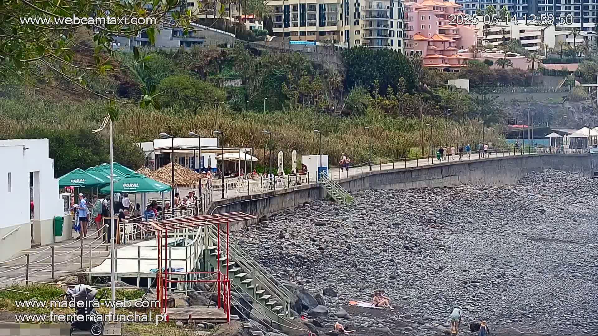 A rocky beach with a paved walkway, buildings, and several people are visible on a sunny day.