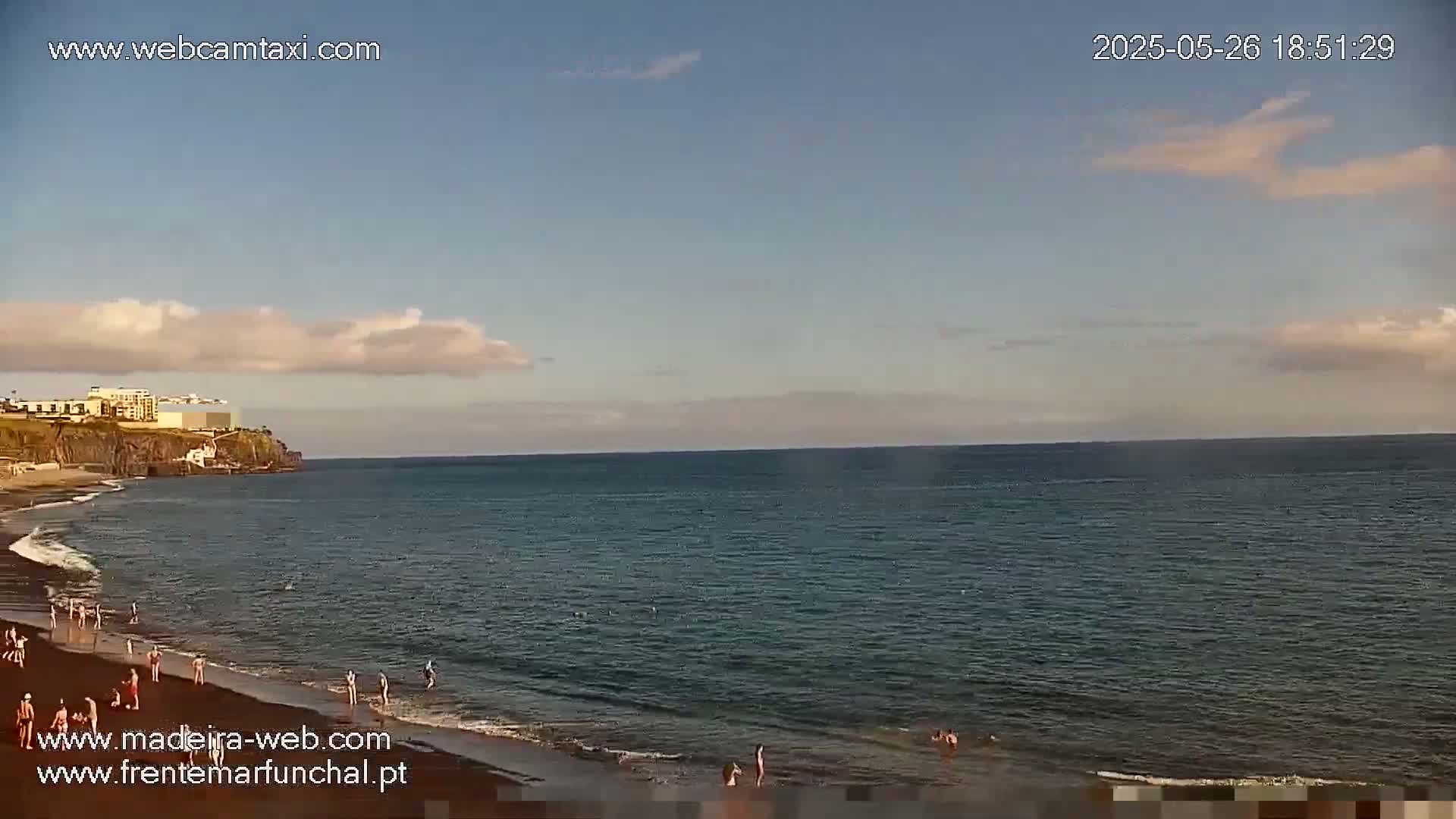 A dark-sand beach with a few people on it stretches along a calm ocean under a partly cloudy sky.