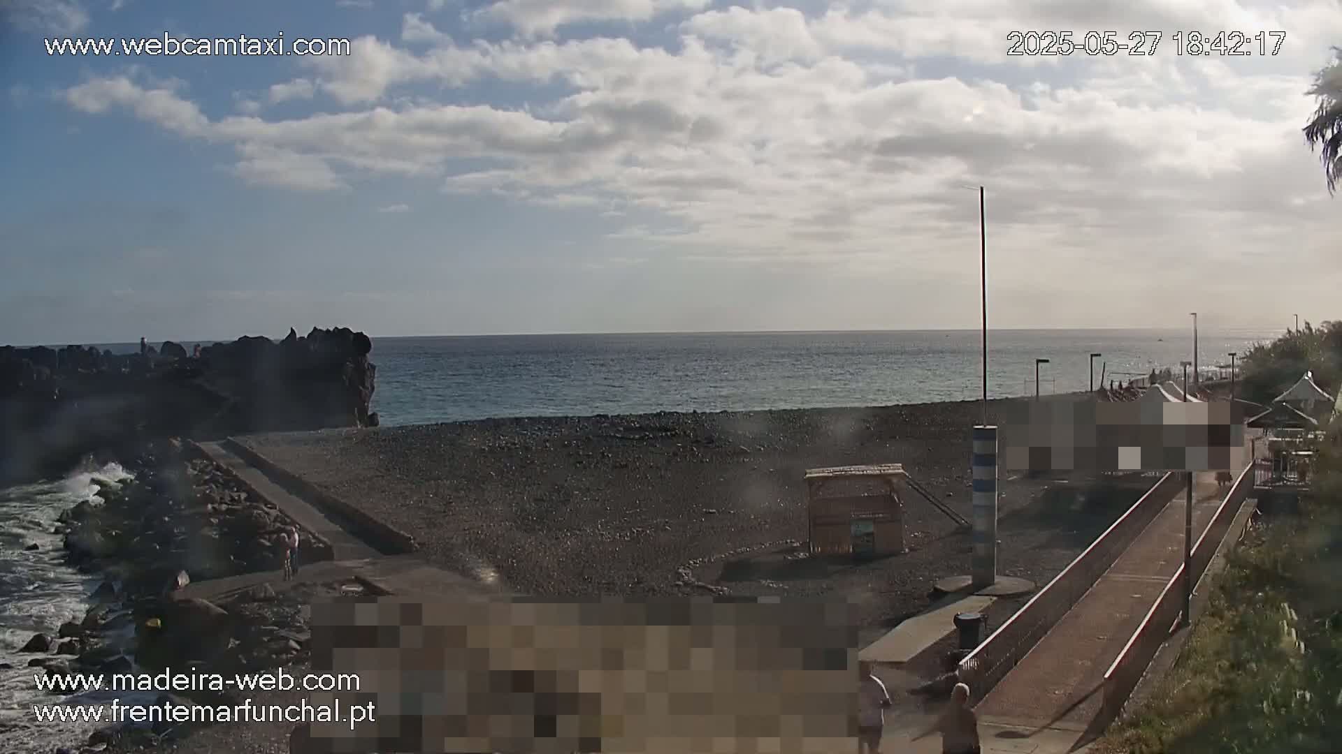 A partly cloudy day reveals a dark-sand beach with a rocky outcrop, a calm ocean, and a paved walkway leading to some structures near the water.