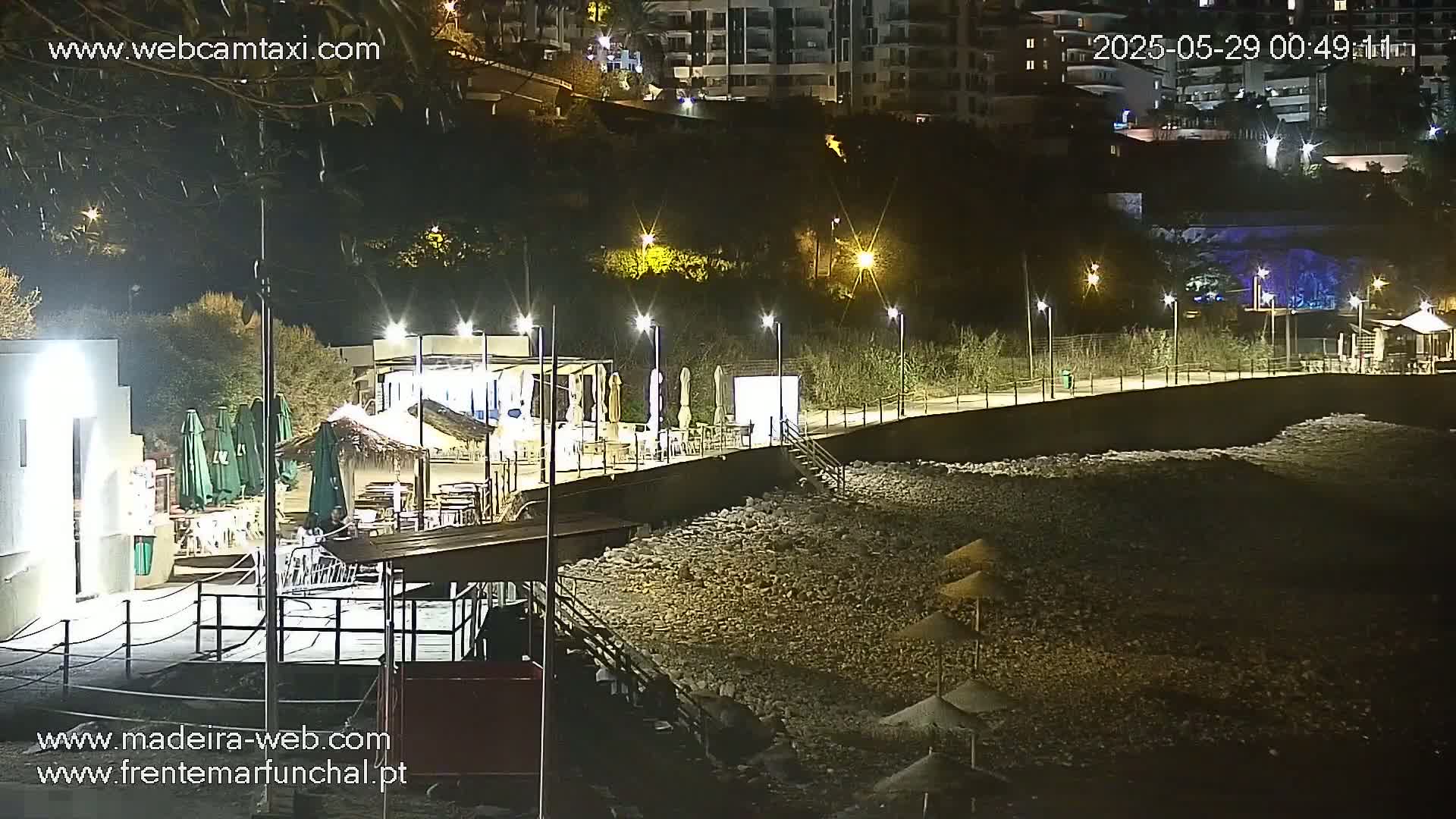 A nighttime view of a rocky beach with a boardwalk lined with outdoor seating areas and lights, adjacent to buildings on a hill.