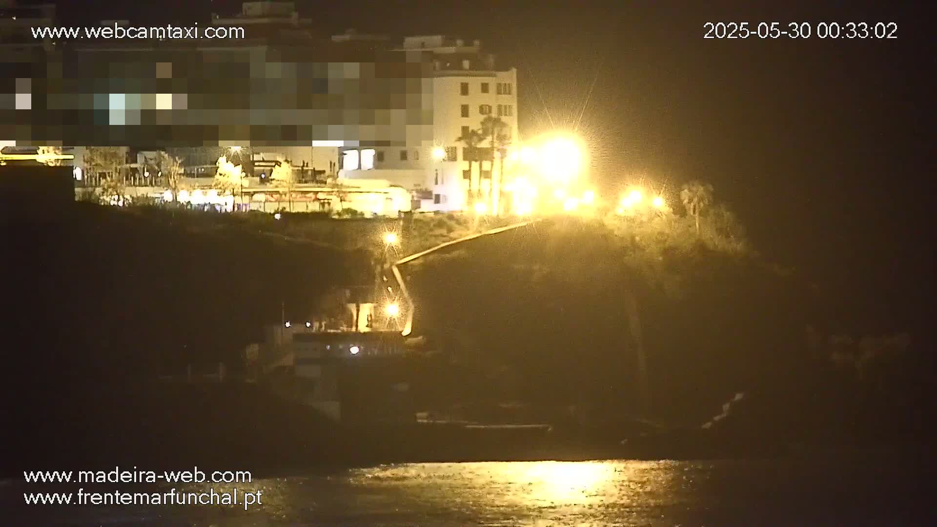A nighttime view of a coastal area shows a building illuminated by streetlights, with calm water reflecting the lights in the foreground.