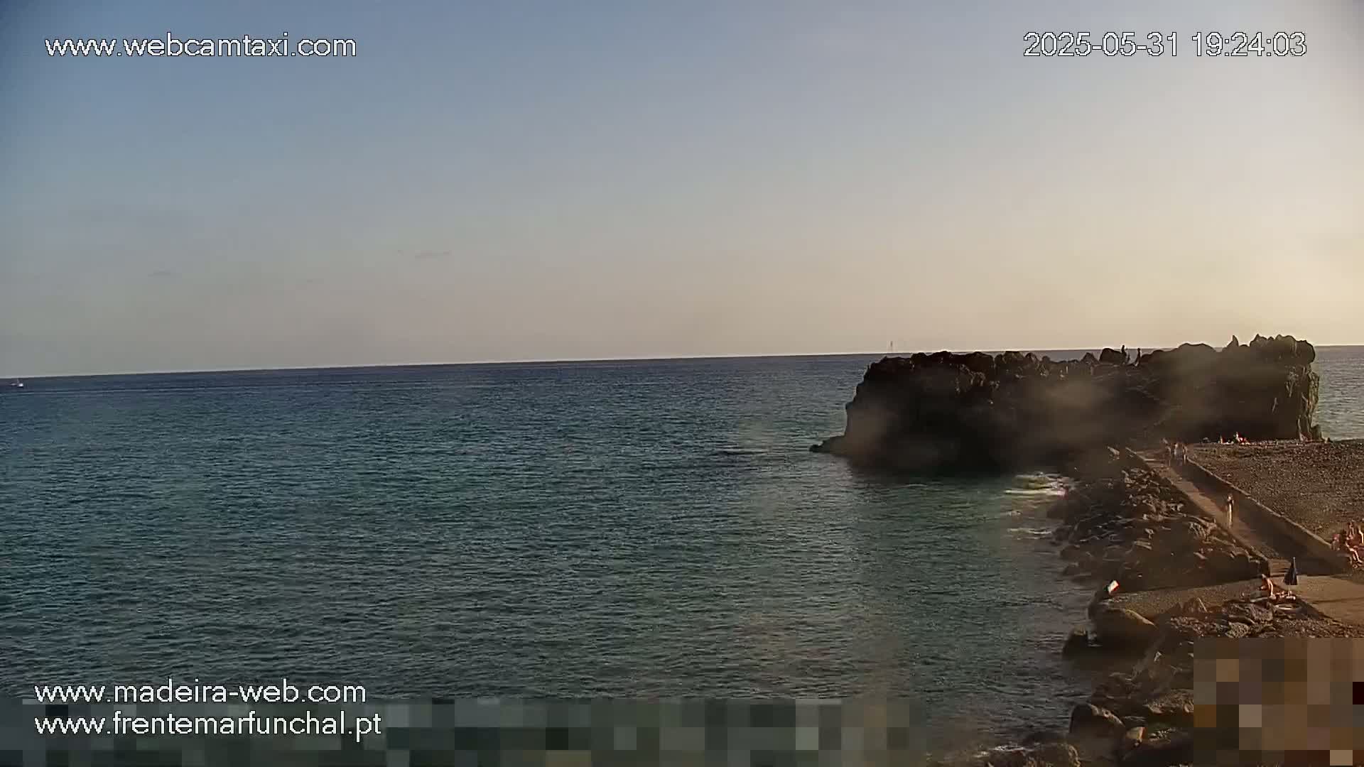 A calm ocean scene shows a rocky outcropping and a paved walkway extending from the shore under a clear, pale sky.