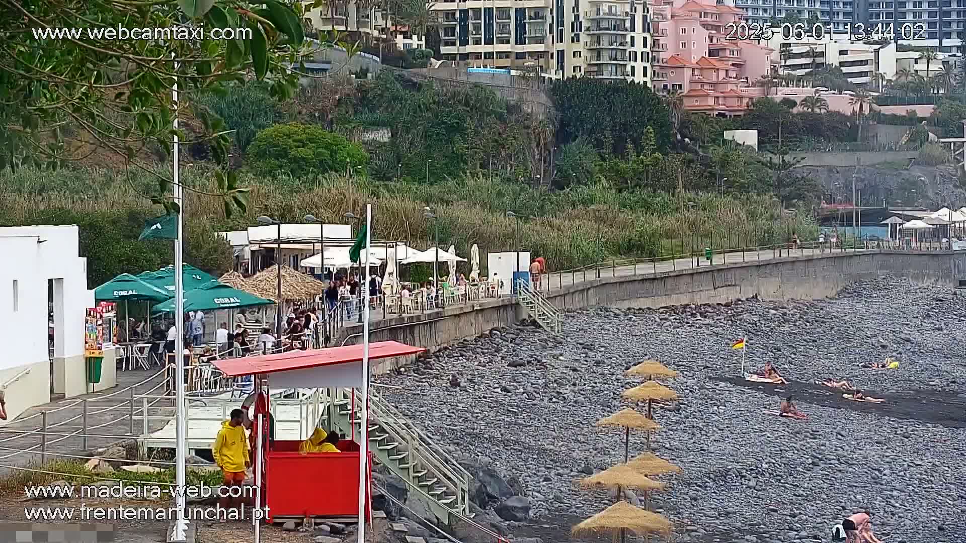 A rocky beach with people sunbathing under umbrellas, near a boardwalk lined with restaurants and buildings, on an overcast day.