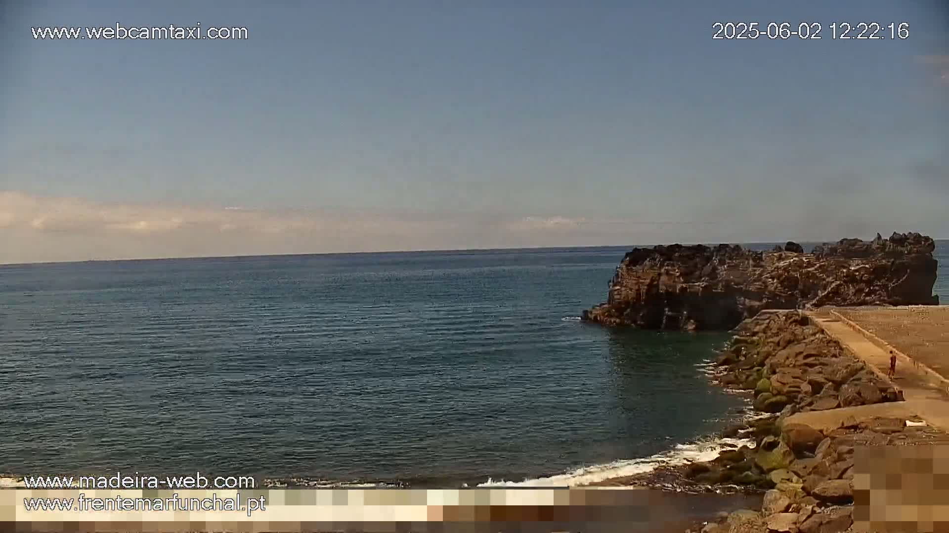 A rocky outcrop juts into a calm, dark-blue ocean under a mostly clear sky.
