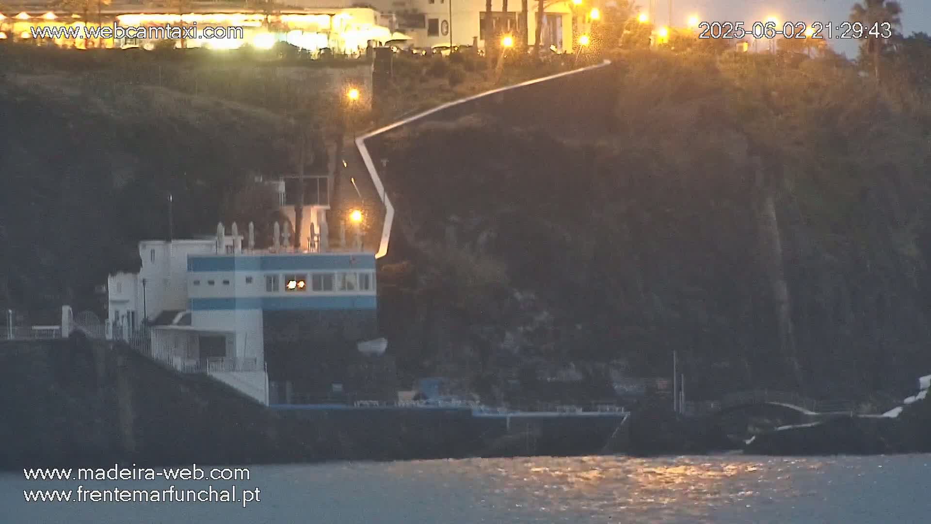 A low-light coastal scene shows a building near the water's edge, set against a dark cliff, under a twilight sky.