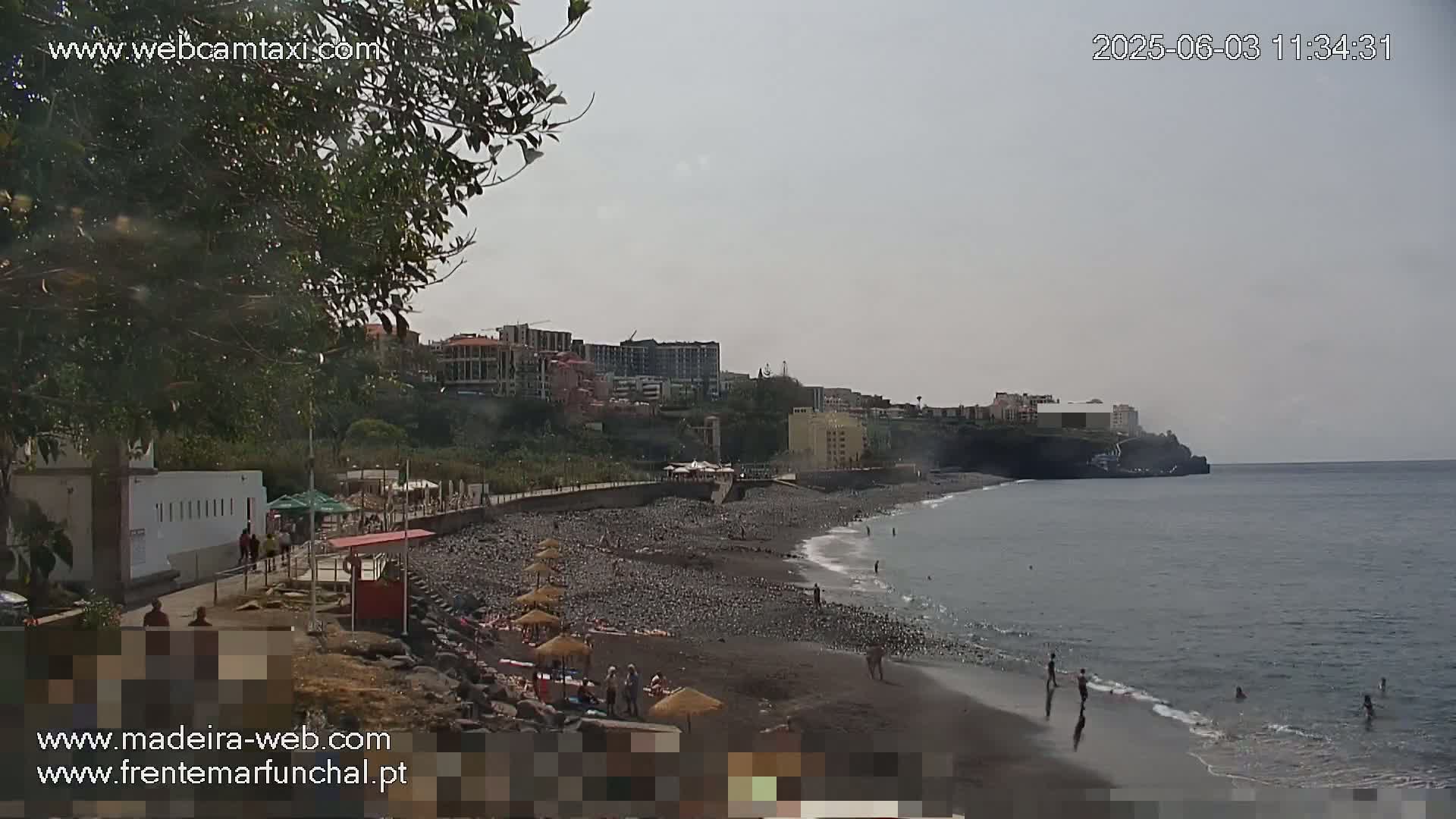 A dark-colored, pebbled beach with a few people on it and buildings on a hillside overlooking a calm ocean under an overcast sky.