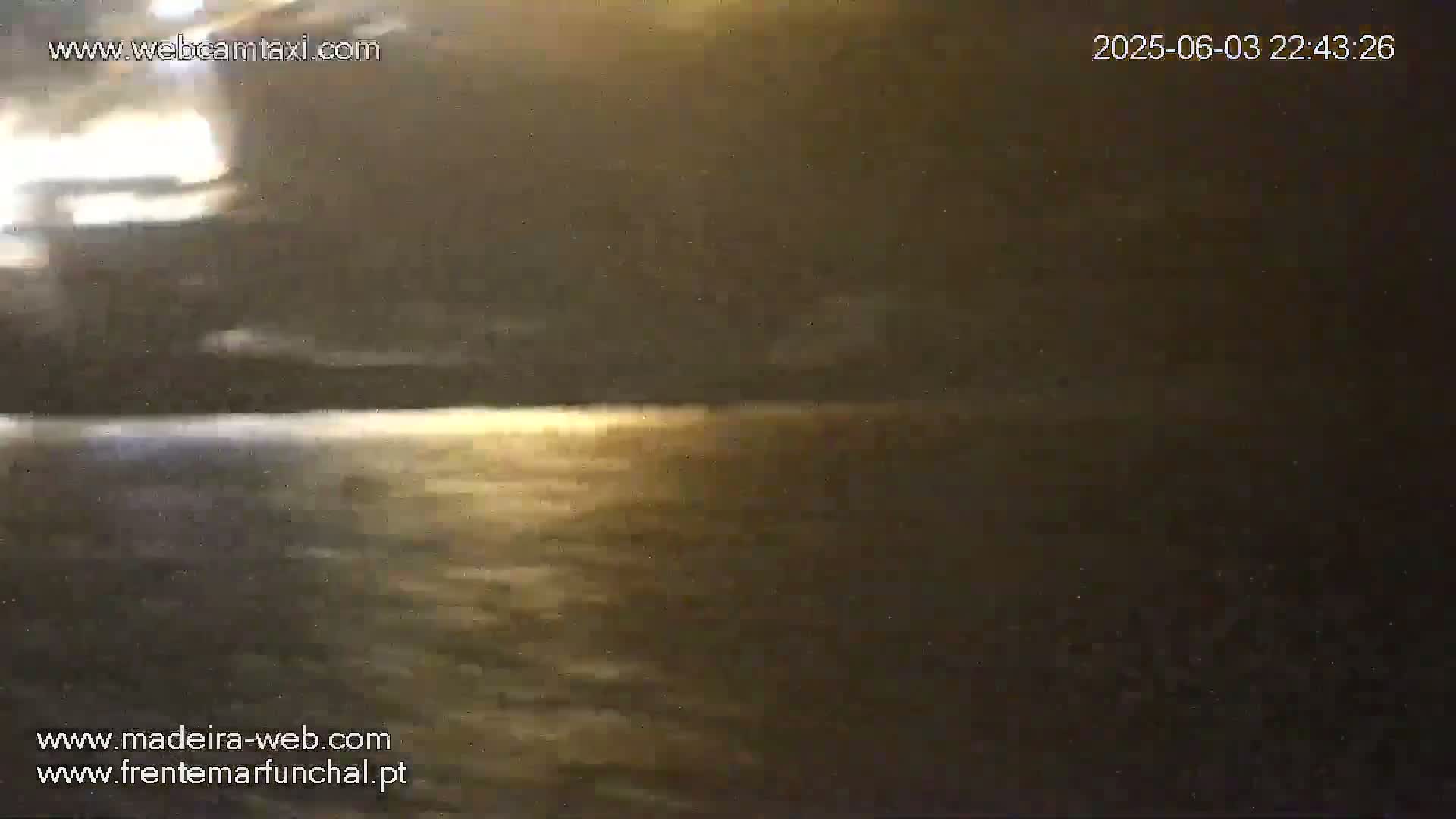 A dark, nighttime view of a calm body of water reflecting a faint light source in the distance under a mostly cloudy sky.