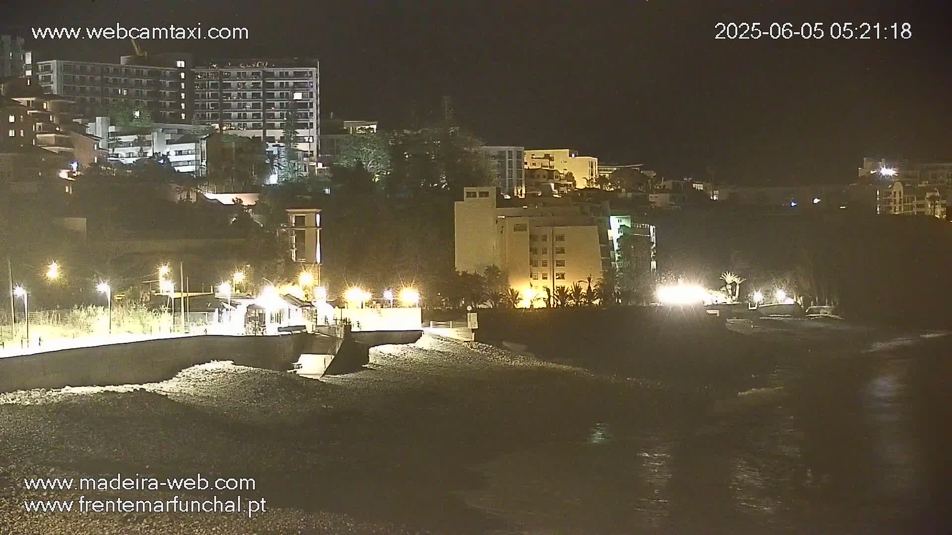 A nighttime view of a rocky beach with dimly lit buildings on a hillside above, and gentle waves lapping the shore.