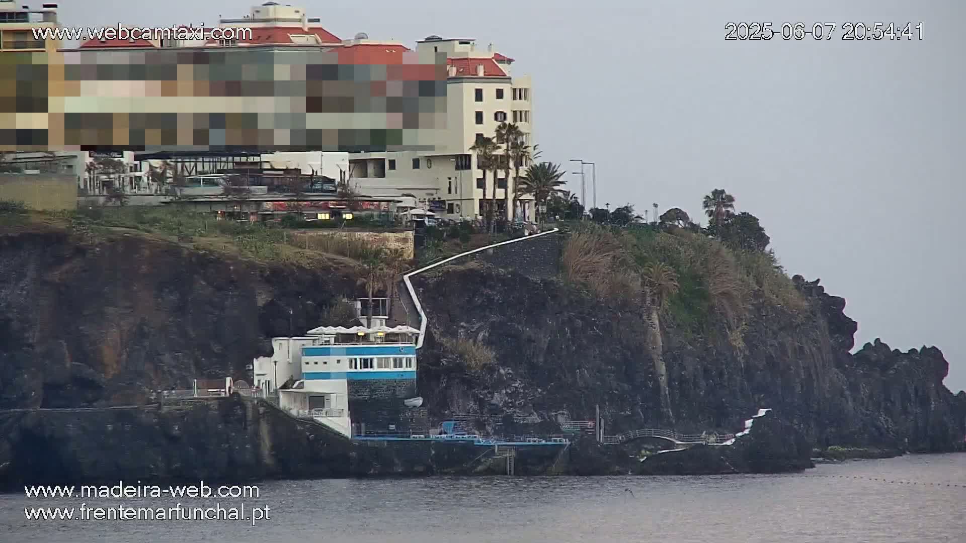 A seaside building complex situated on a dark cliff overlooking calm water under an overcast sky.