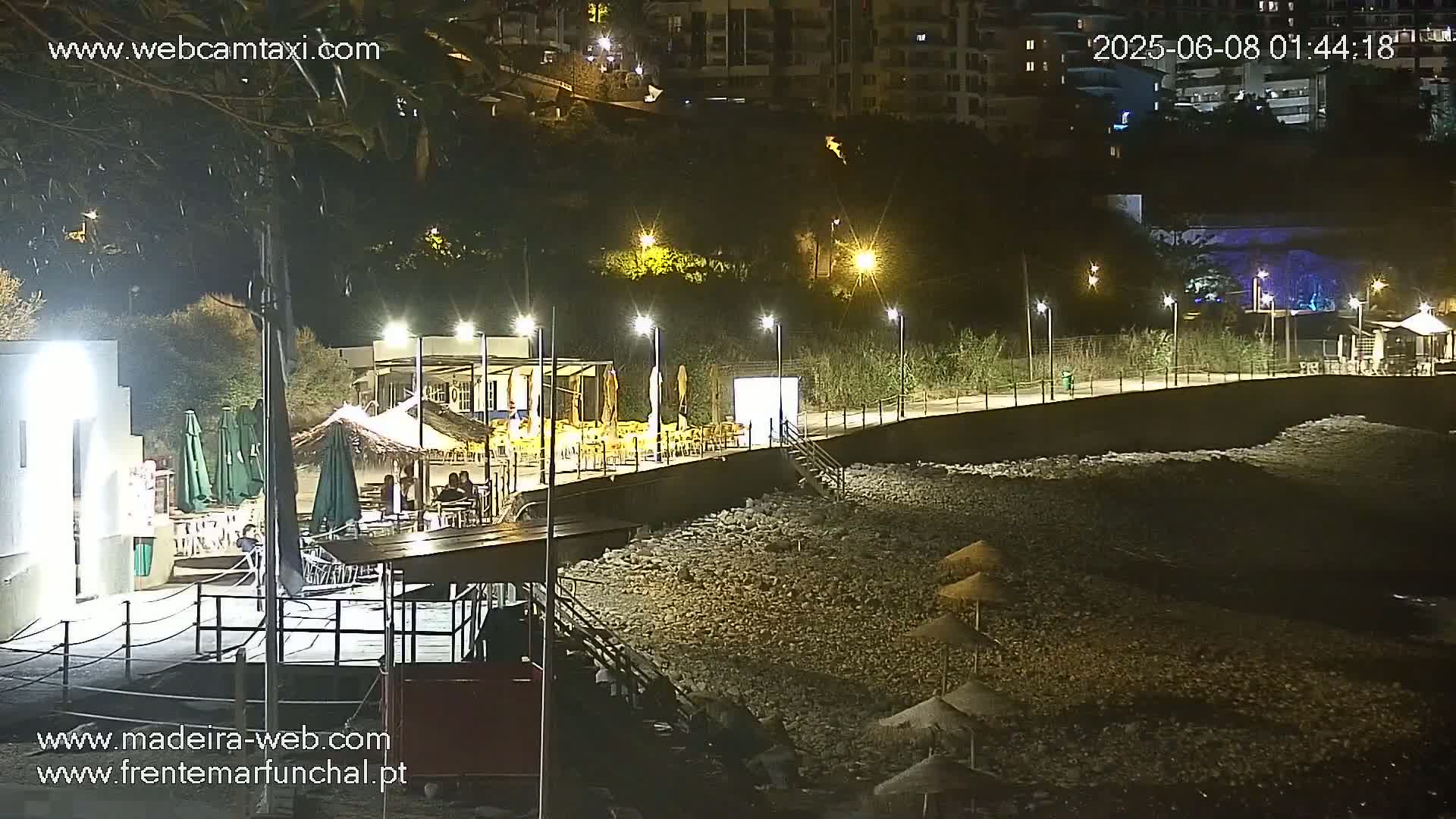A nighttime view of a pebble beach with outdoor seating areas and lighting, adjacent to buildings on a hillside.