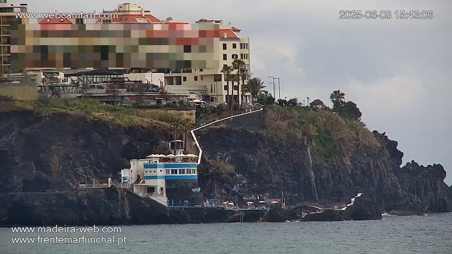 A partially obscured hotel sits atop dark volcanic cliffs overlooking a calm ocean on an overcast day.