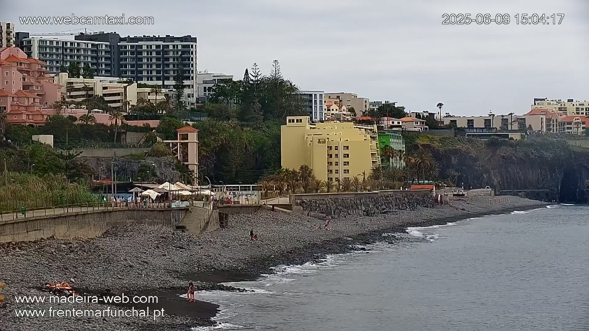 A rocky beach with a few people on it is next to a sea wall and buildings on a hillside under an overcast sky.