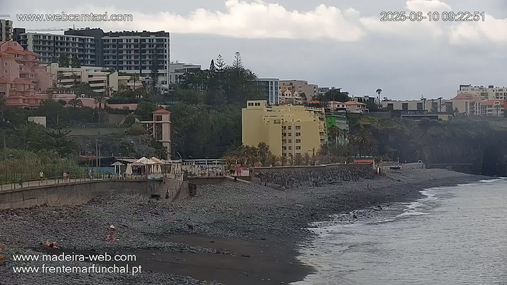 A dark-colored pebble beach stretches along a coastline, with buildings and hotels rising up a hillside under an overcast sky.