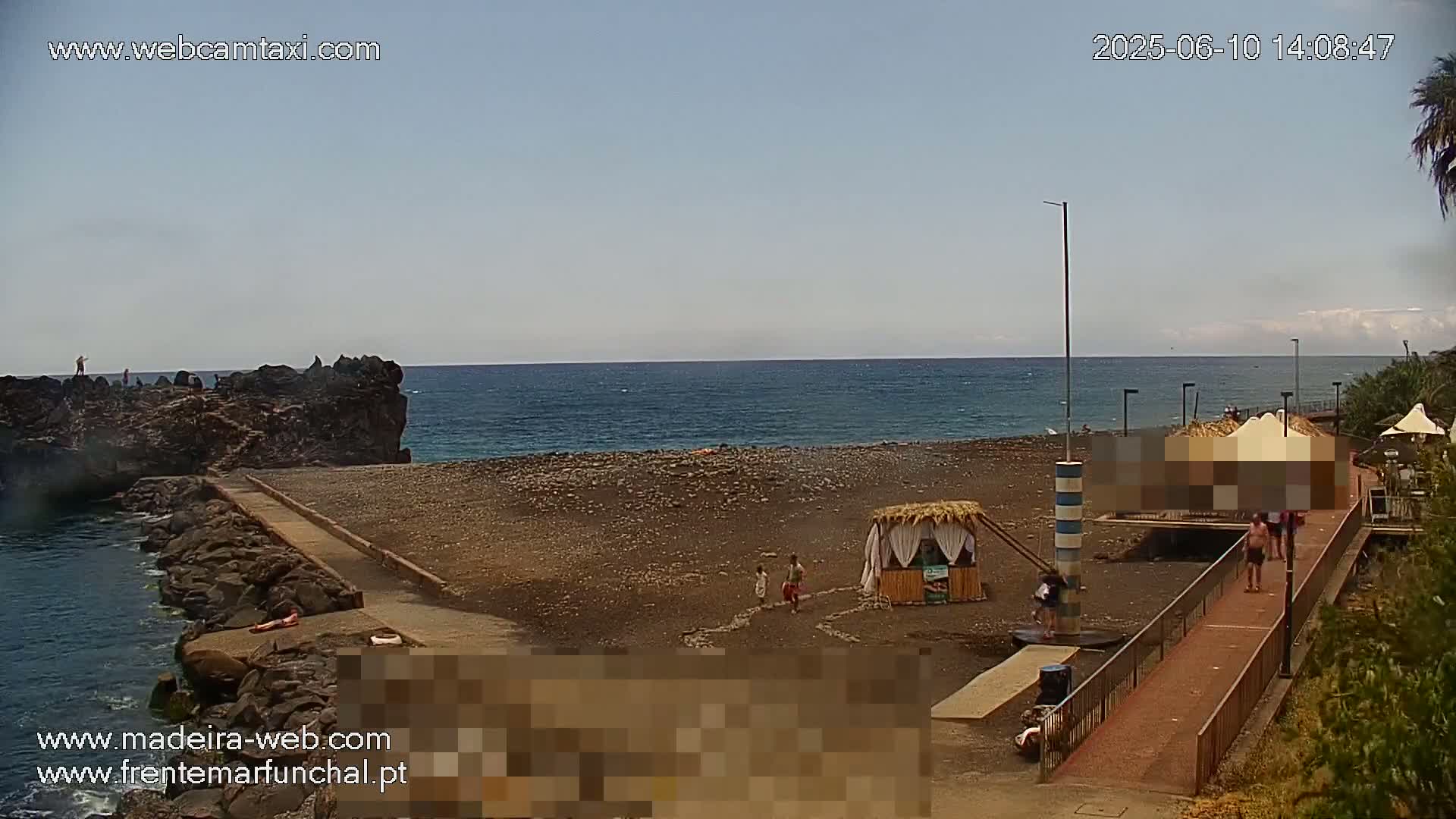 A mostly sunny day at a rocky beach with calm ocean waters, showing a few people on the shore and near a small structure.
