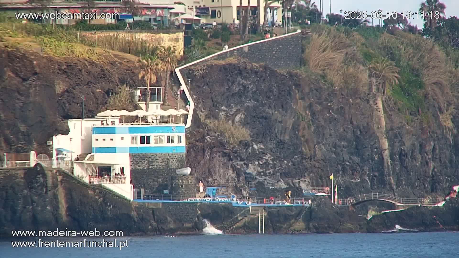 A blue and white building with a patio sits atop dark volcanic rock near the ocean, under a mostly sunny sky.