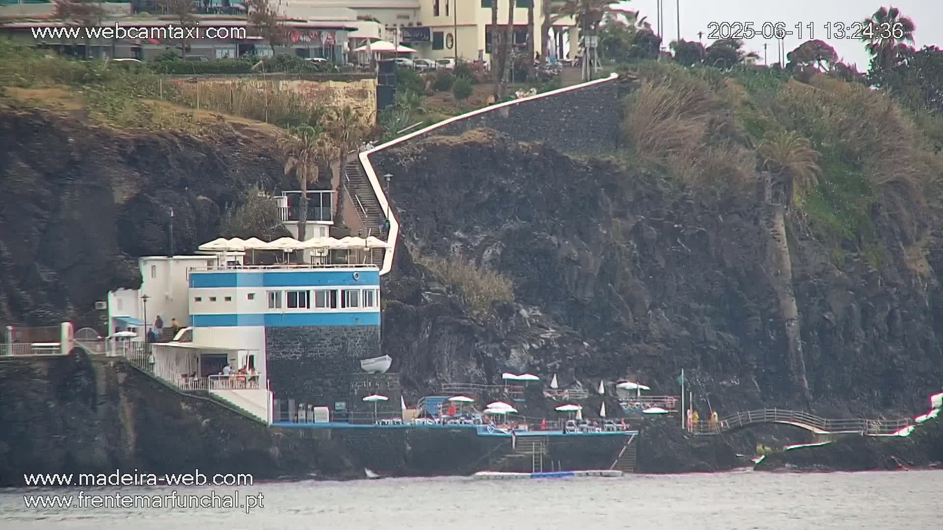 A seaside restaurant with outdoor seating is situated on a rocky cliff overlooking calm water.