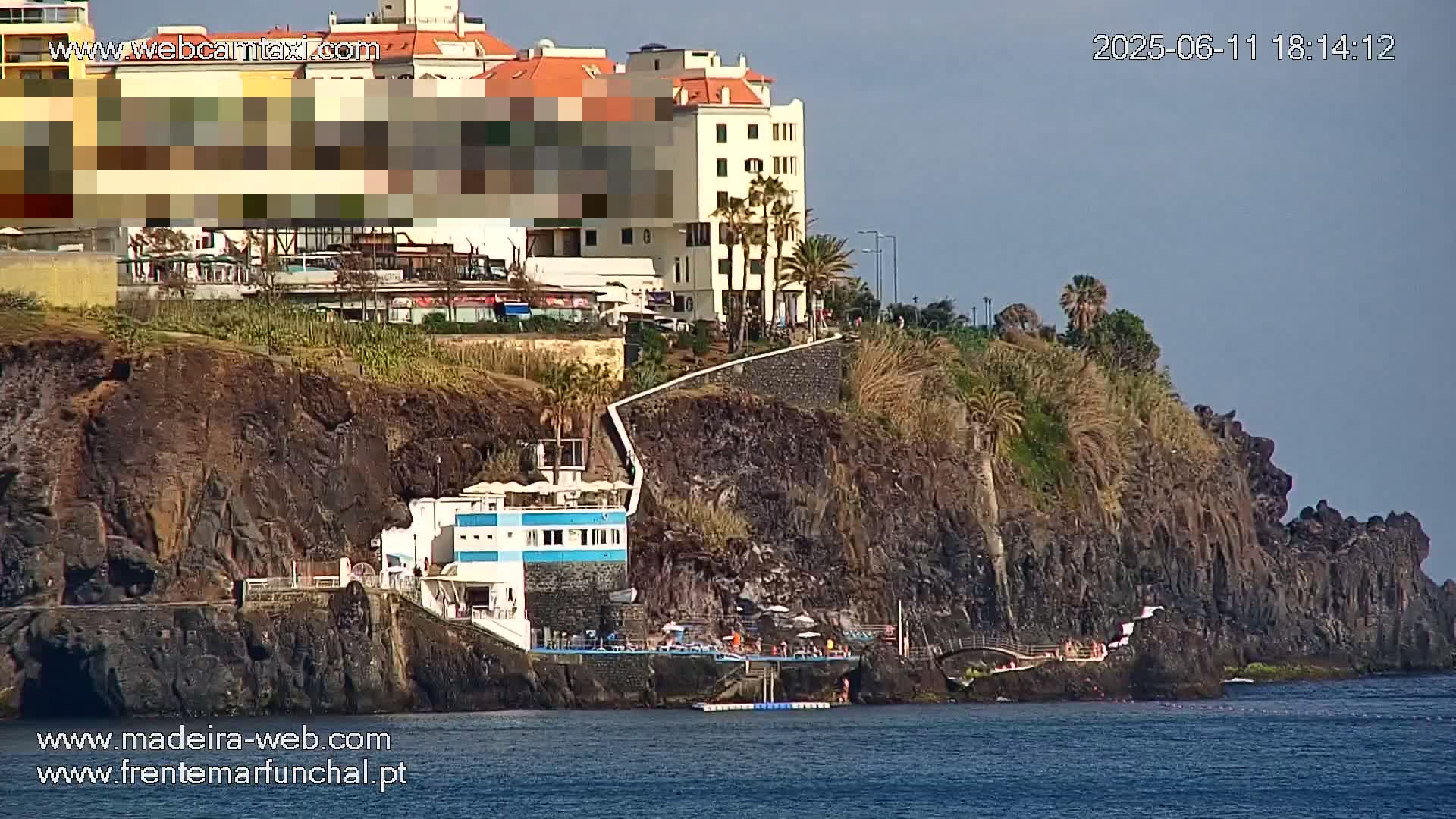 A seaside cliff with buildings and a walkway on top, and a small beach area and building at the bottom, under a clear sky.