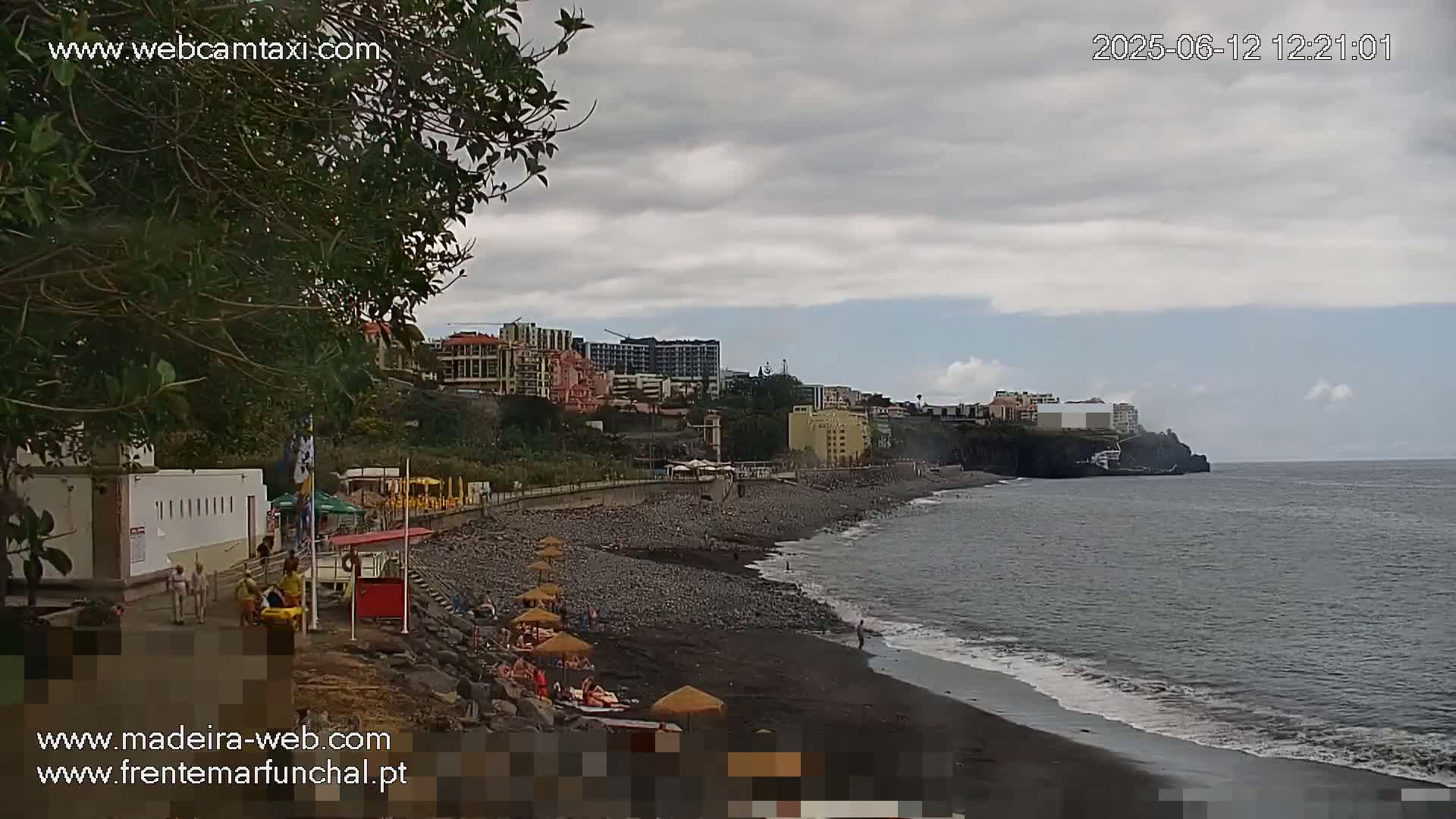 A dark-sand beach with several people and umbrellas is seen next to a coastal town under a mostly cloudy sky.