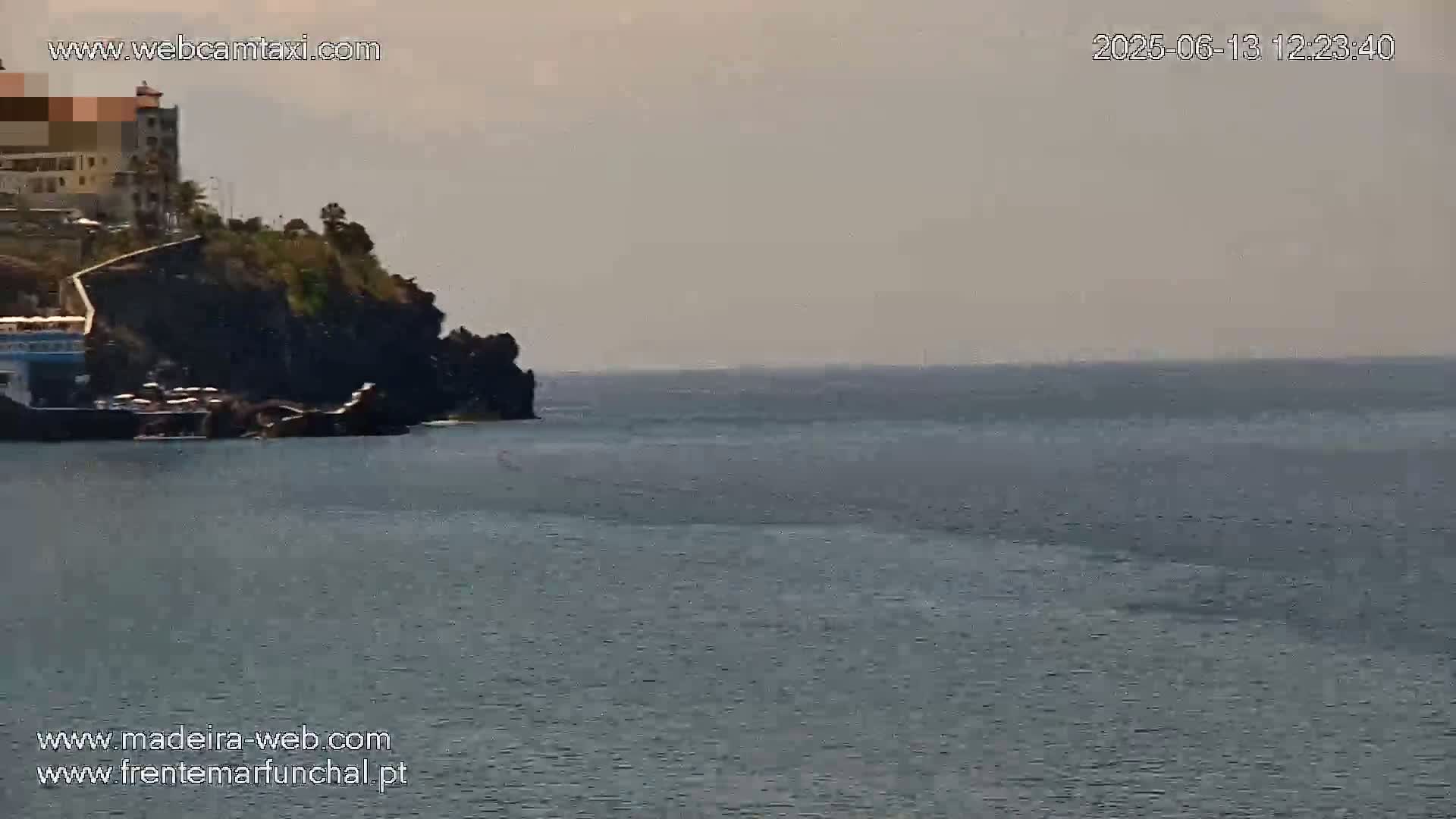 A hazy, calm ocean stretches to the horizon, meeting a rocky shoreline where a building and some vegetation are visible.