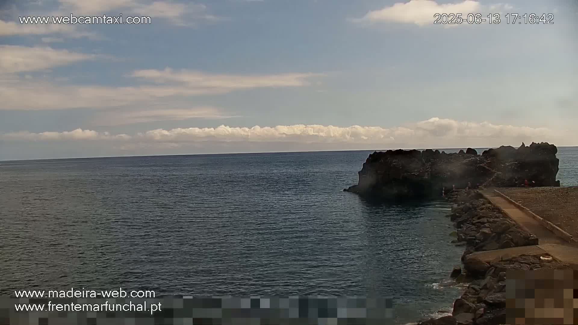 A mostly sunny day shows a dark rocky outcropping jutting into a calm, dark-blue ocean.