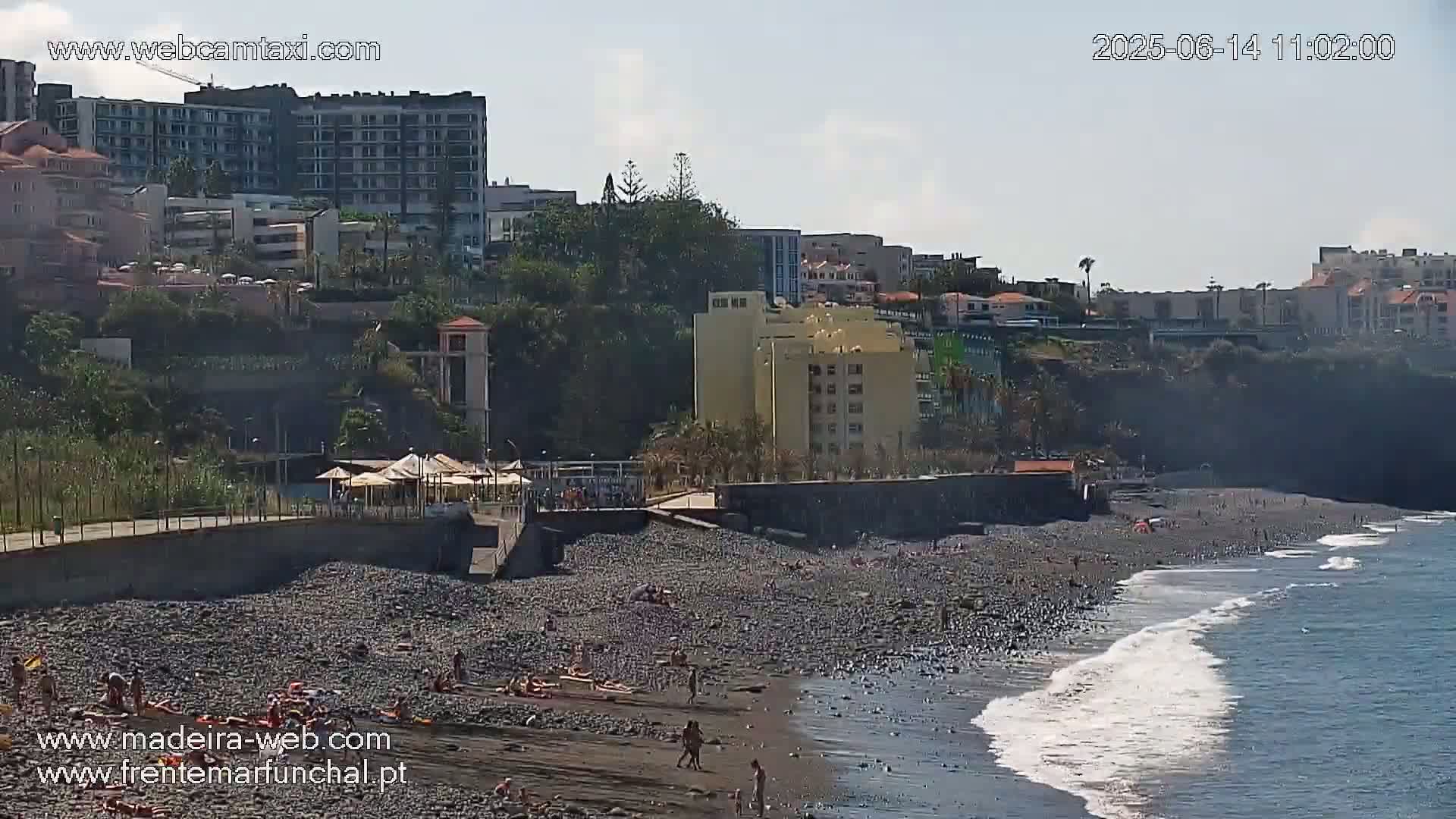 A pebble beach with scattered sunbathers is situated below buildings and cliffs on a sunny day with calm ocean waves.