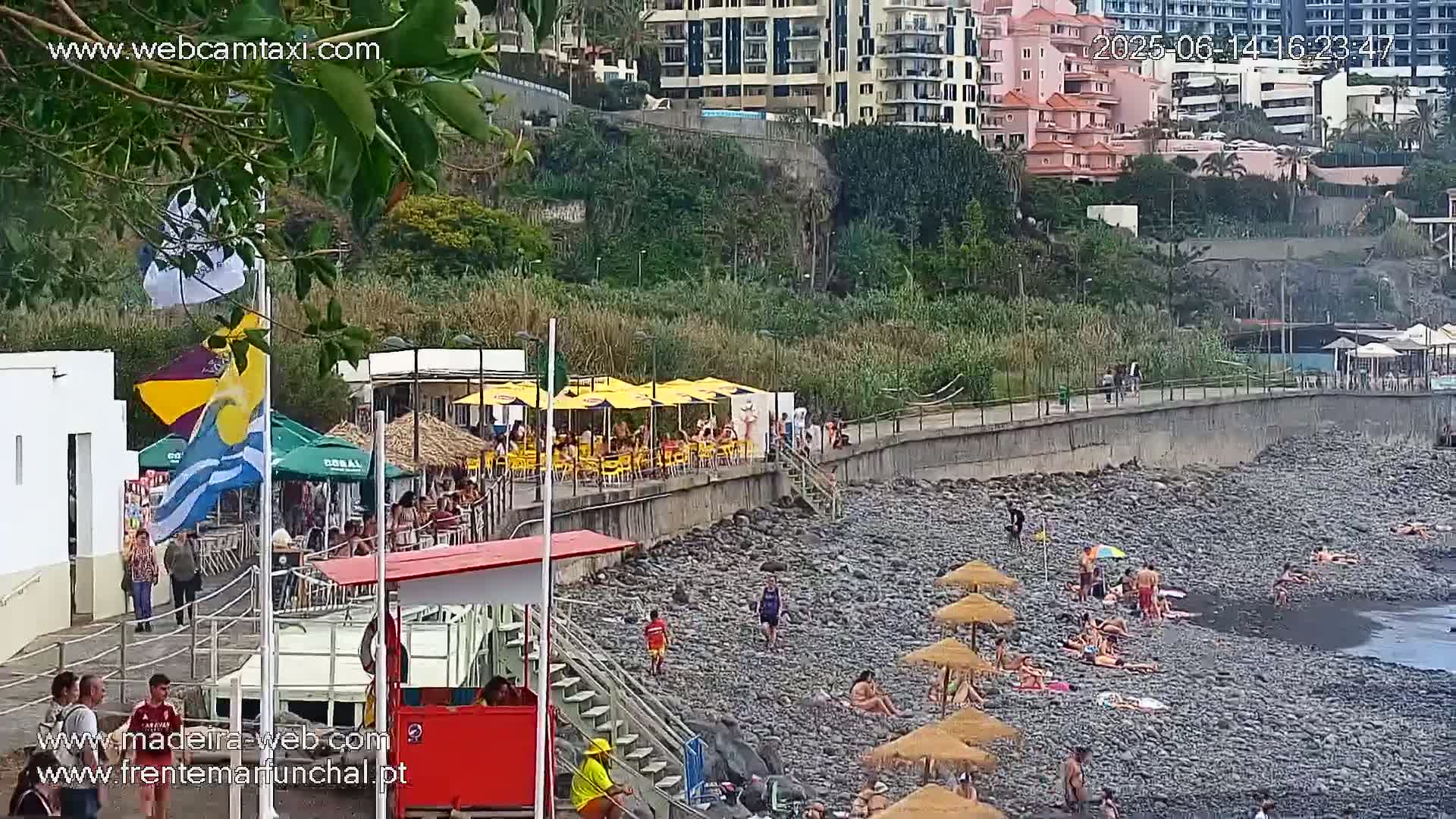 A rocky beach with people sunbathing under umbrellas, near a promenade with restaurants and buildings on a hillside, in overcast weather.