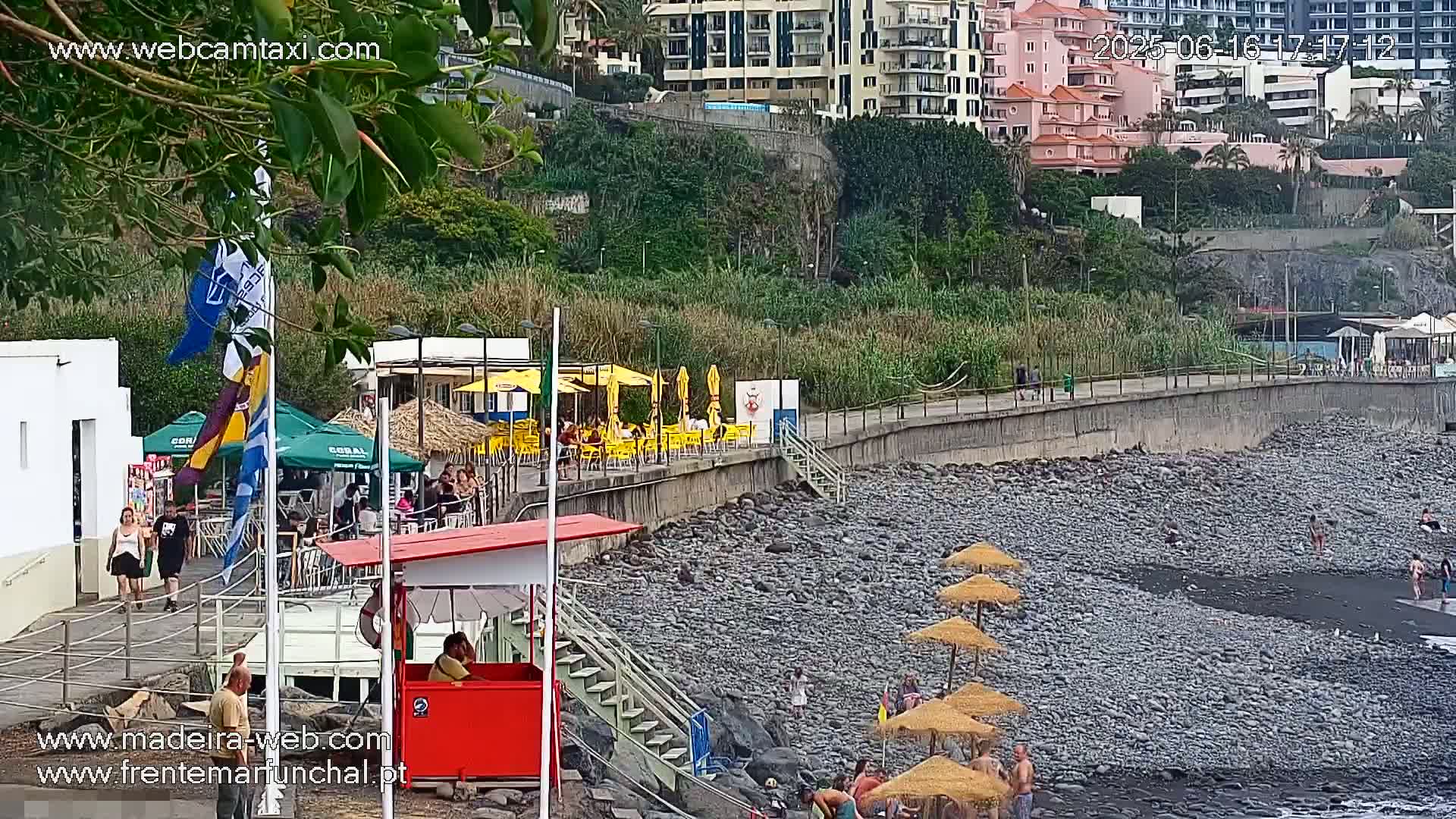 A rocky beach with several people near yellow umbrellas and a red lifeguard stand next to a building and a boardwalk, partially obscured by trees.