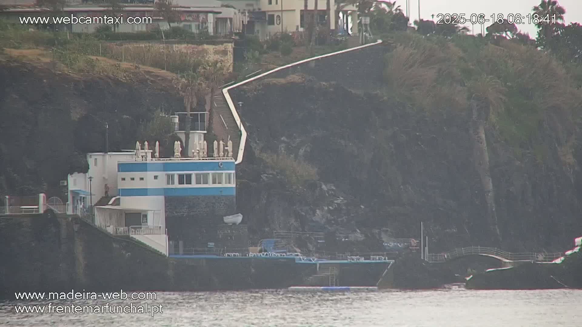 A blue and white building sits on a dark, rocky cliff overlooking calm water, with a long walkway leading up to buildings on the hillside above.