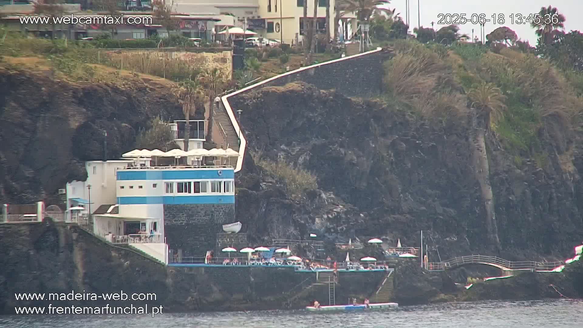 A seaside building with a blue stripe and a sunbathing area on a rocky cliff overlooks calm water.