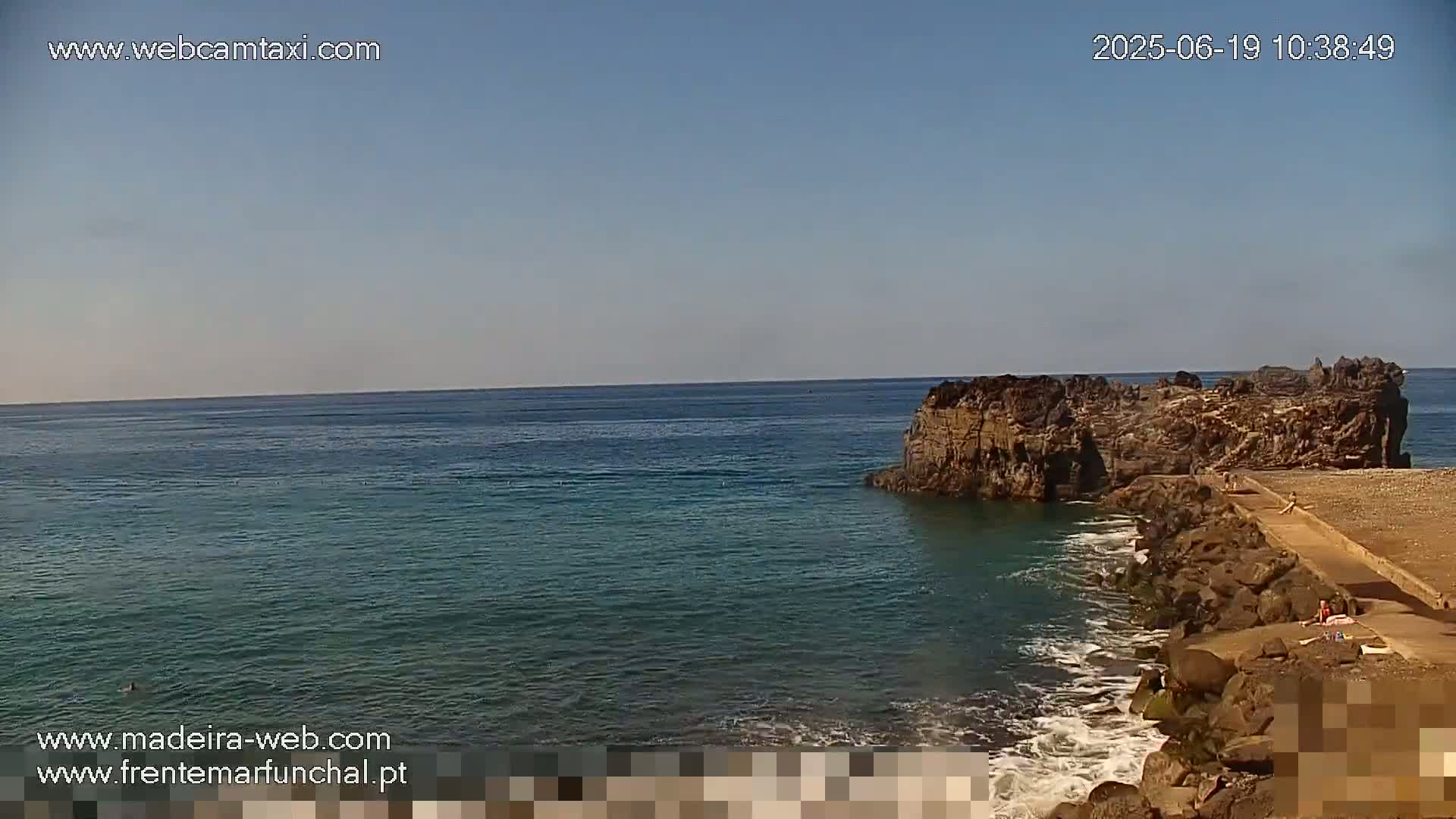 A rocky outcrop juts into a calm, clear ocean under a bright, sunny sky.