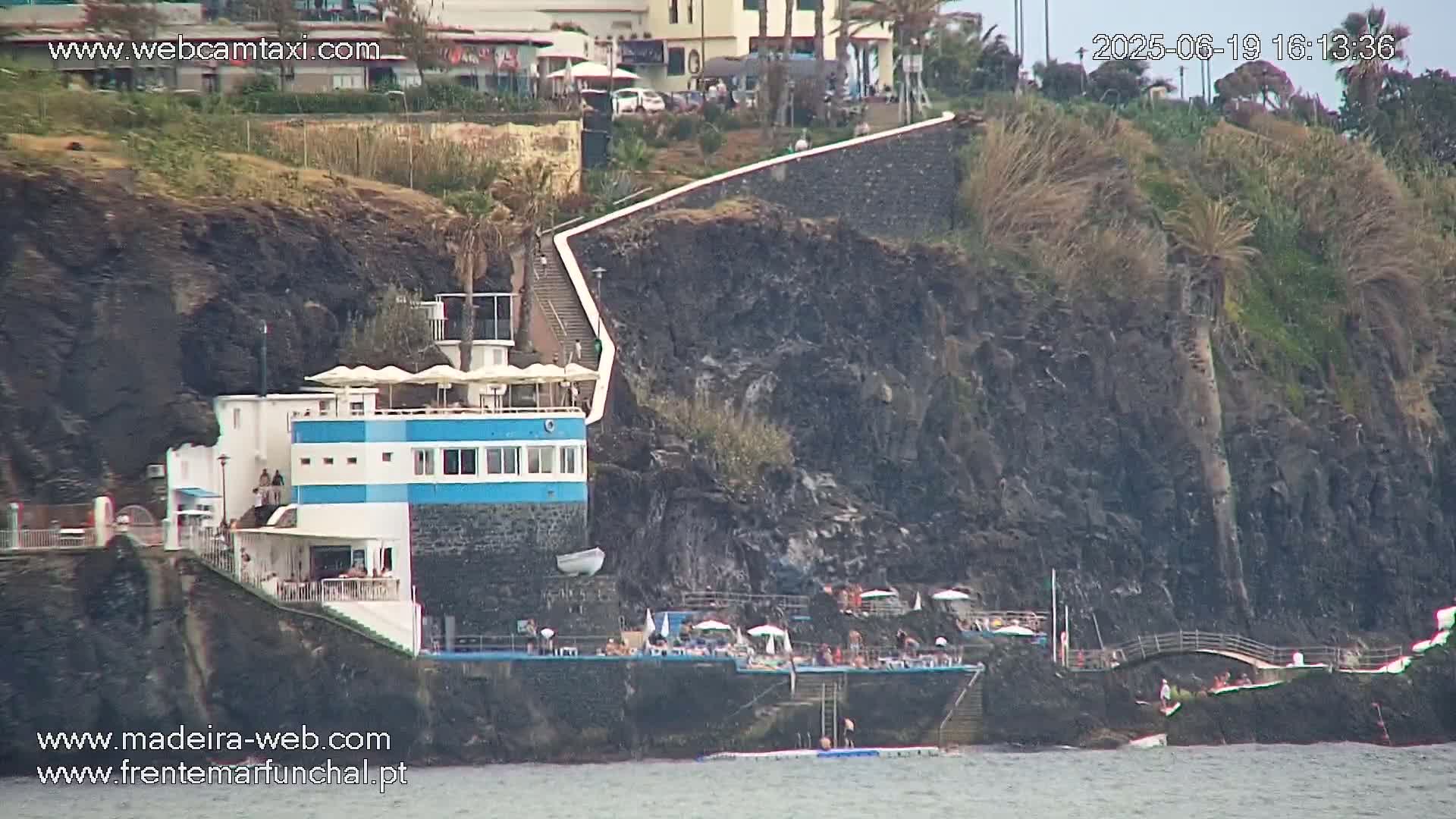 A seaside building with a blue stripe and outdoor patio area is situated on a dark, rocky cliff overlooking calm water.