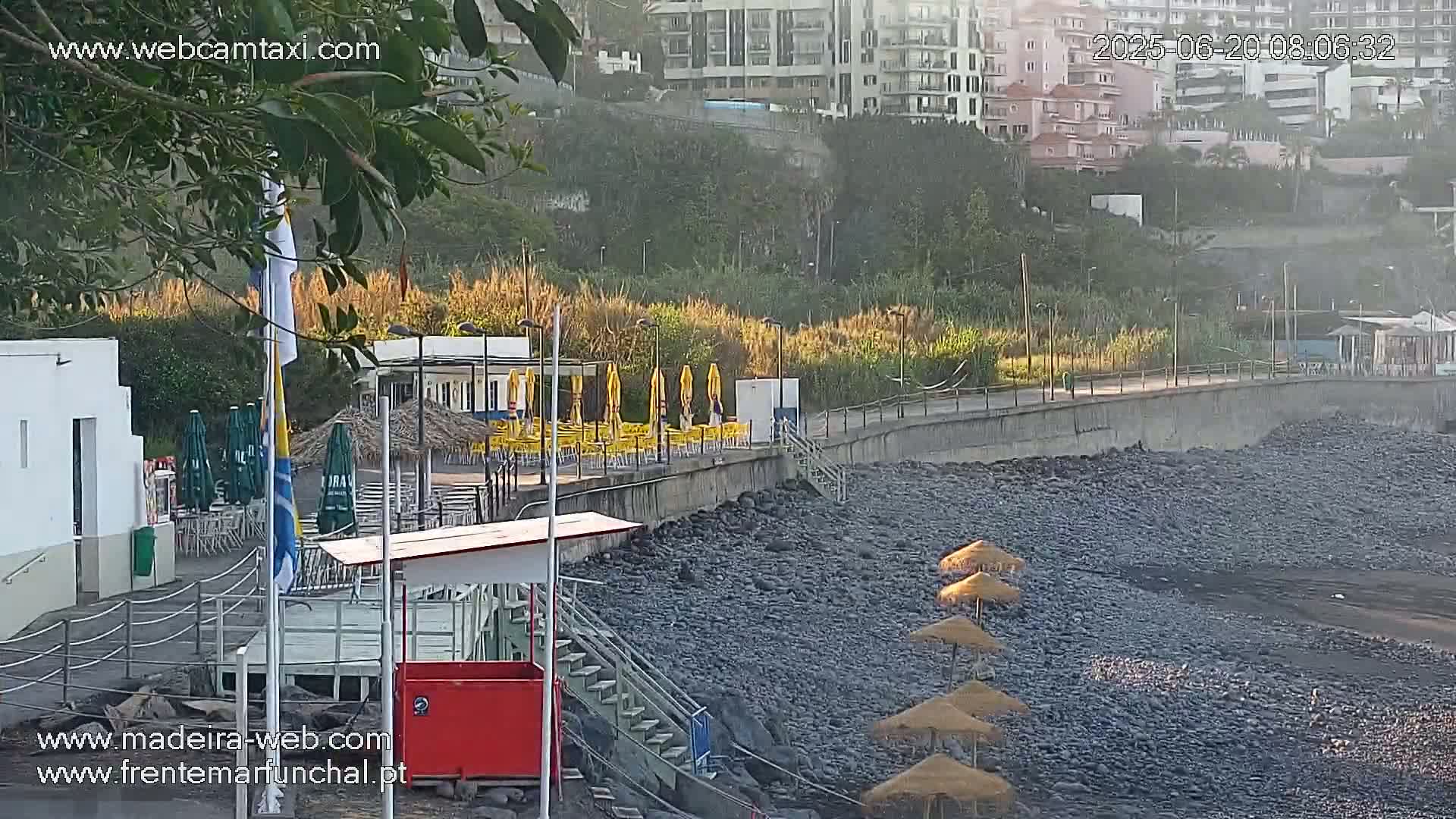 A rocky beach with several straw umbrellas and yellow patio umbrellas is situated below a concrete wall and buildings, partially obscured by hazy morning air.