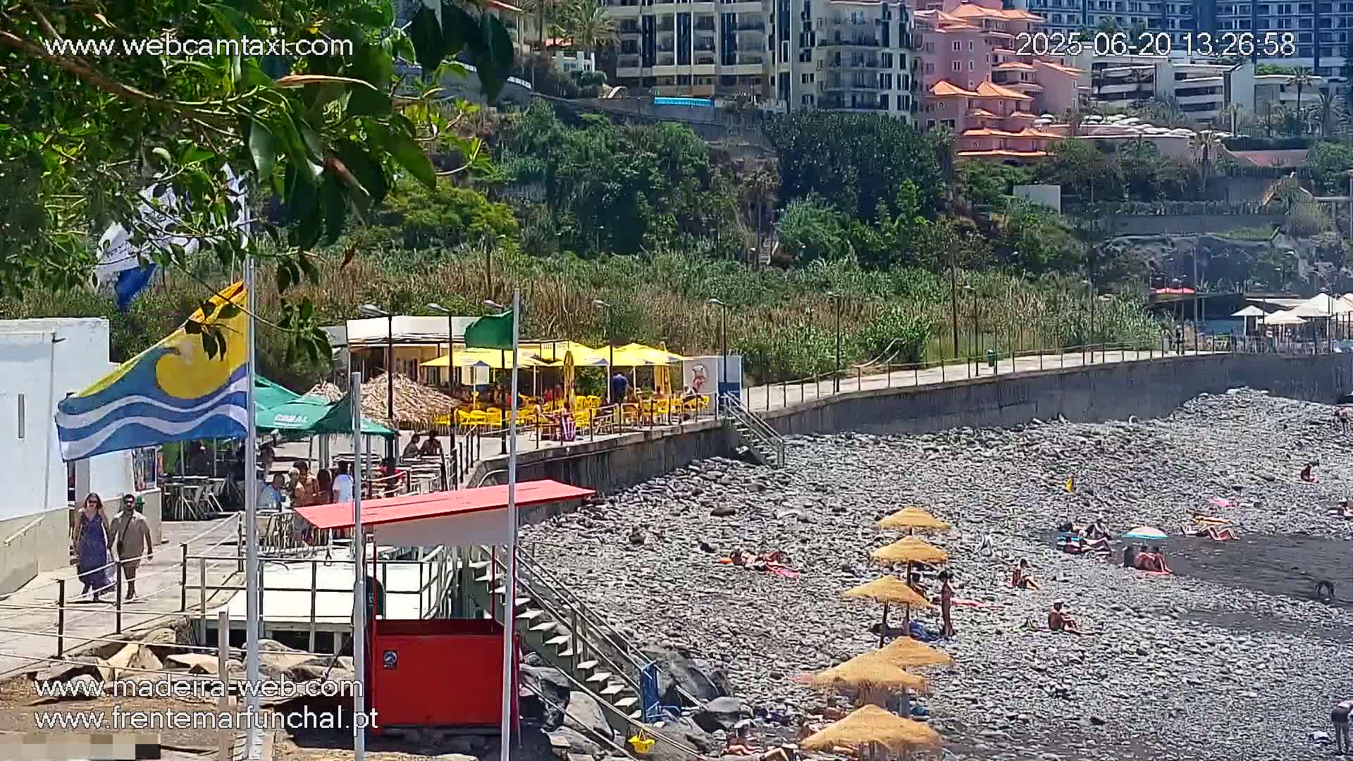 A rocky beach with people sunbathing under straw umbrellas, a boardwalk with cafes, and buildings on a hillside in sunny weather.