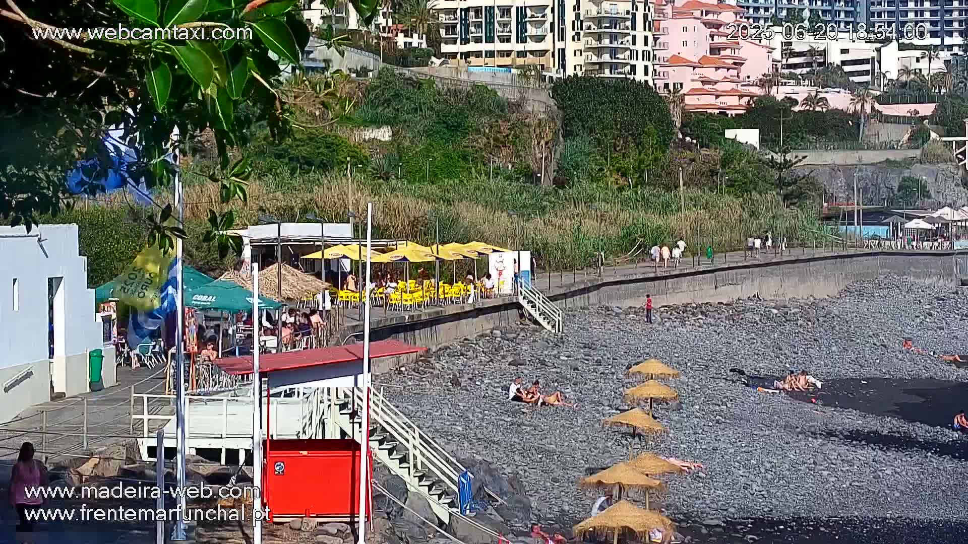 A rocky beach with people sunbathing under umbrellas, near a boardwalk lined with restaurants and buildings, on a sunny day.