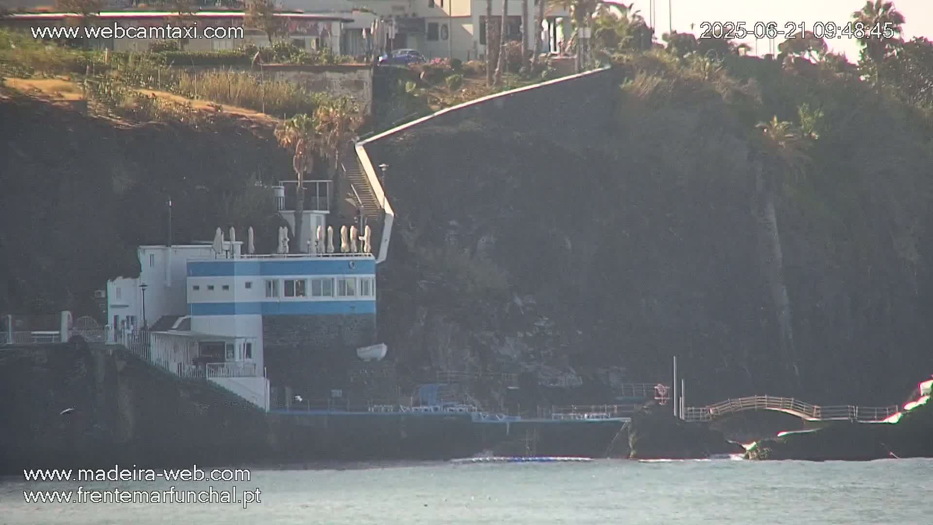 A blue and white building is situated on a rocky cliff overlooking a calm body of water, with a small bridge connecting to a nearby rocky outcrop under sunny conditions.