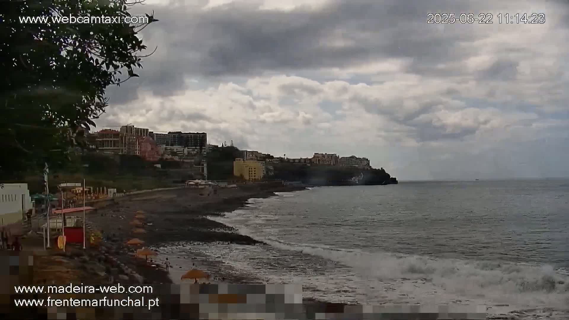 A dark-sand beach with a few people on it is next to buildings on a hillside overlooking a calm ocean under a cloudy sky.
