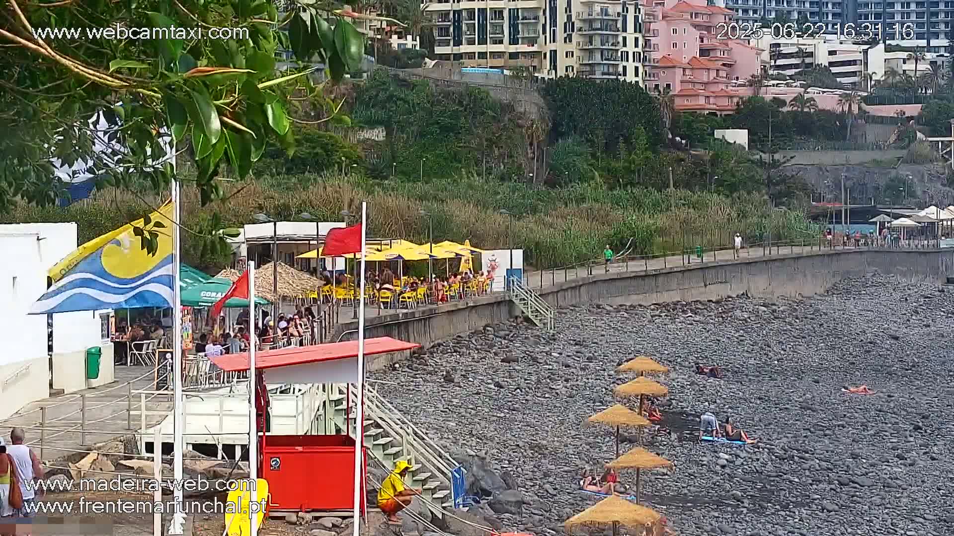A rocky beach with people sunbathing under umbrellas, a lifeguard station, and an outdoor restaurant with yellow umbrellas sits below a walkway and buildings on a sunny day.