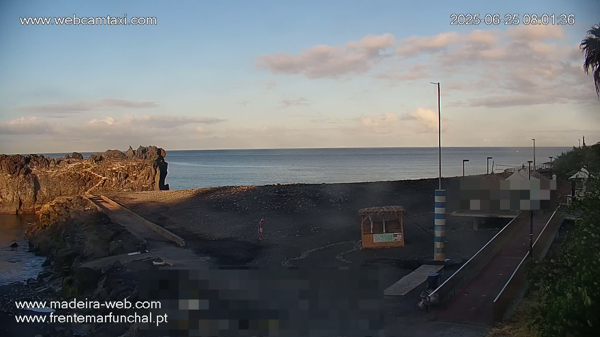 A mostly clear sky overlooks a dark-sand beach with a rocky outcropping and a small wooden structure near a walkway leading to a partially obscured building.